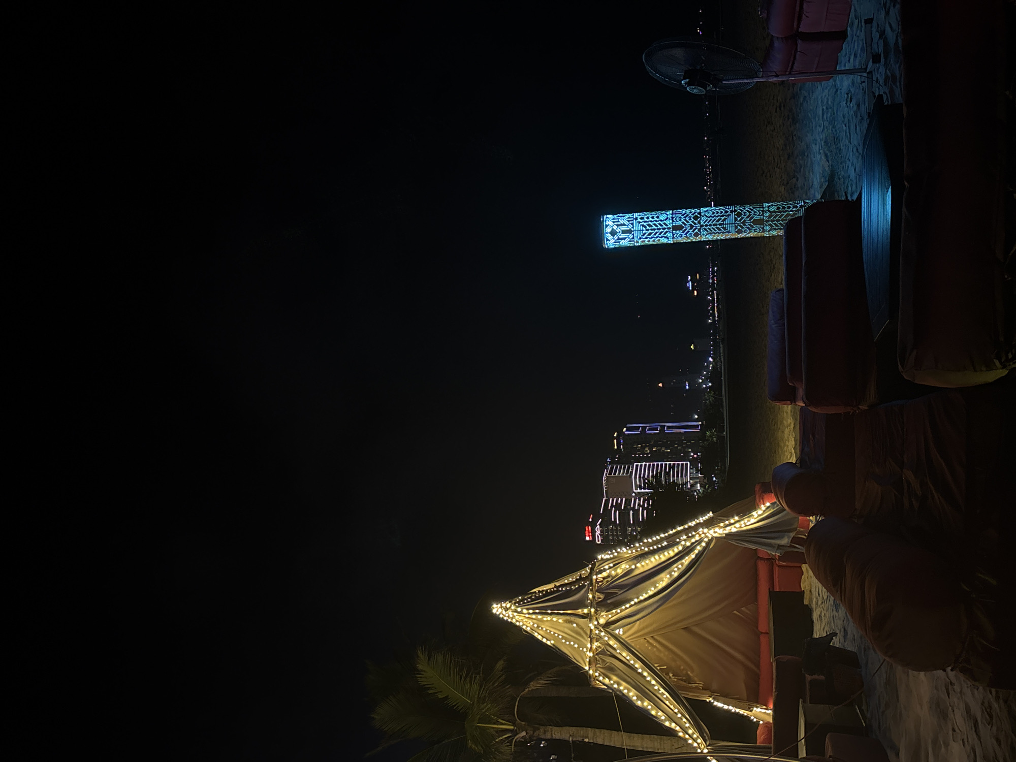 This is a nighttime scene on a beach in Đà Nẵng, Vietnam. A decorated tent-like structure adorned with string lights is partially visible at the bottom. Palm trees are visible in the lower left corner. In the background, the city skyline is illuminated, with prominent buildings and lights. A tall structure with patterned blue lighting is also visible. Lounge chairs and a standing fan are present on the beach.