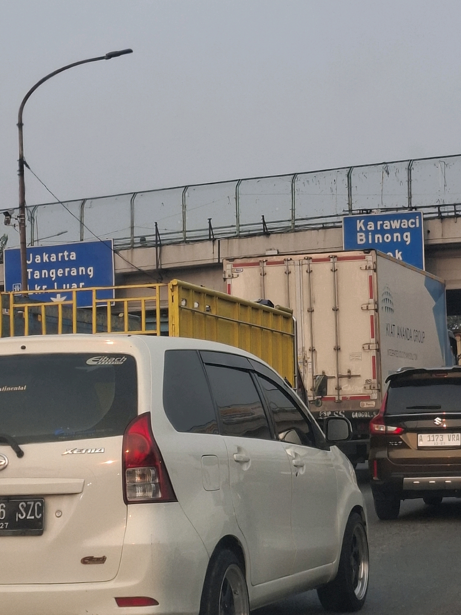 This is a medium-angle, slightly low-shot, outdoor, daytime photograph of a traffic jam on a highway in Tangerang, Indonesia. The sky is overcast. In the foreground, a white multi-purpose vehicle is partially visible on the left. To its right, a dark brown SUV is also partially visible. Behind the vehicles, a yellow truck is seen, followed by a white semi-trailer truck. Overhead, a blue highway sign indicates directions to "Jakarta Tangerang" and "Karawaci Binong". The image captures a typical scene of heavy traffic congestion on Indonesian roads.