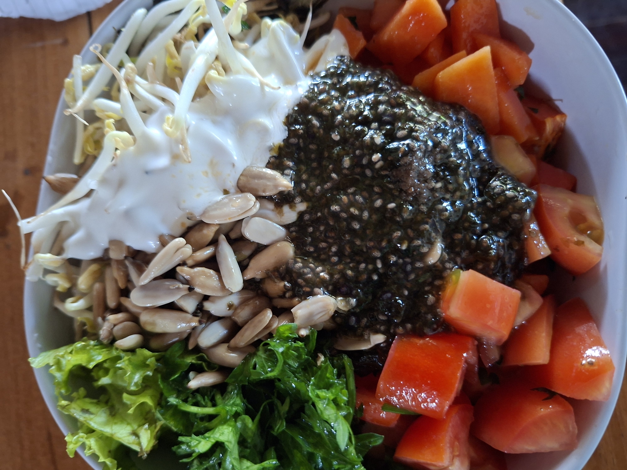 The image shows a bowl of salad. The ingredients include bean sprouts with white creamy sauce, sunflower seeds, chia seeds, diced tomatoes, papaya chunks, and two types of lettuce. The photograph was taken in Lodtunduh, Indonesia.