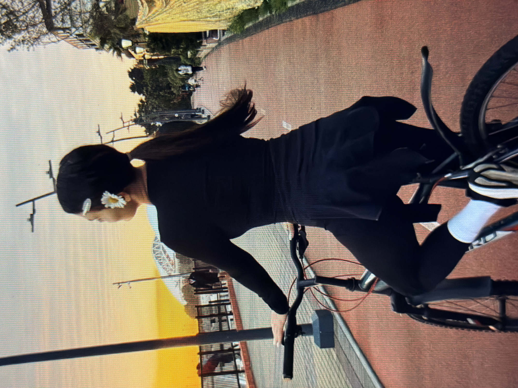 A woman, from behind, rides a bicycle along a paved path in Sirius, Russia. The woman has dark hair pulled back in a ponytail, with a white flower in her hair. She is wearing black leggings, a black long-sleeved shirt, and a black skirt or overlay. Her right hand is on the bicycle handlebars. The bicycle is dark colored with red accents. The path is reddish-orange and curves to the right. There are streetlights and trees lining the path on the right. In the background, there is a body of water, a bridge, and a yellow structure. The sky is a gradient of orange and light blue, indicating it is either sunrise or sunset. Other people are visible in the background, some walking and some on bicycles.