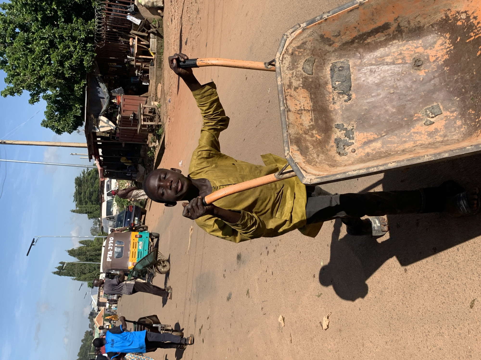 In Gura Top, Nigeria, a young boy pushes a wheelbarrow on a dirt road. The boy wears a long-sleeved, mustard-colored tunic and dark pants. The wheelbarrow is orange and weathered, showing signs of heavy use. In the background, other people are visible, some walking and others near a small shop or market stall. A motorized tricycle with "PTA 3970" written on it is parked on the road. The scene appears to be a bustling urban or semi-urban environment, with a few trees and buildings in the distance. The sun casts long shadows, indicating it is likely late afternoon.