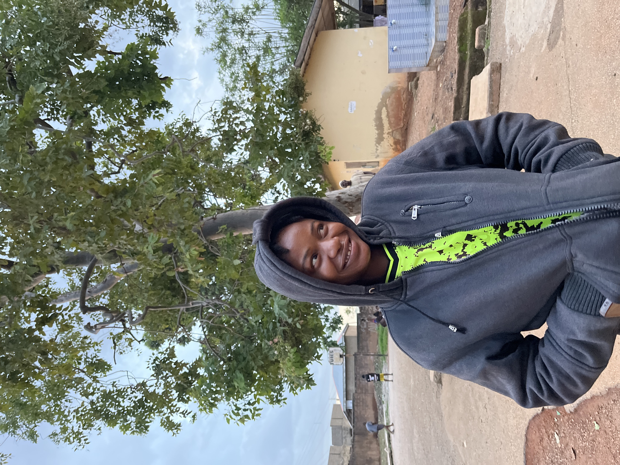 In Jos, Nigeria, a young woman with dark skin and dark hair, wearing a gray hooded jacket over a neon green shirt, stands in the foreground. She is smiling and looking to her right. Behind her, a dirt court is visible, with a basketball hoop and backboard mounted on a pole. Two young boys are playing basketball in the background. Further back, a small, tan building with a brown roof is situated, and a man stands in the doorway. To the right of the building, a blue tiled water fountain with two spigots is present. The environment is an outdoor urban setting with trees and a cloudy sky.