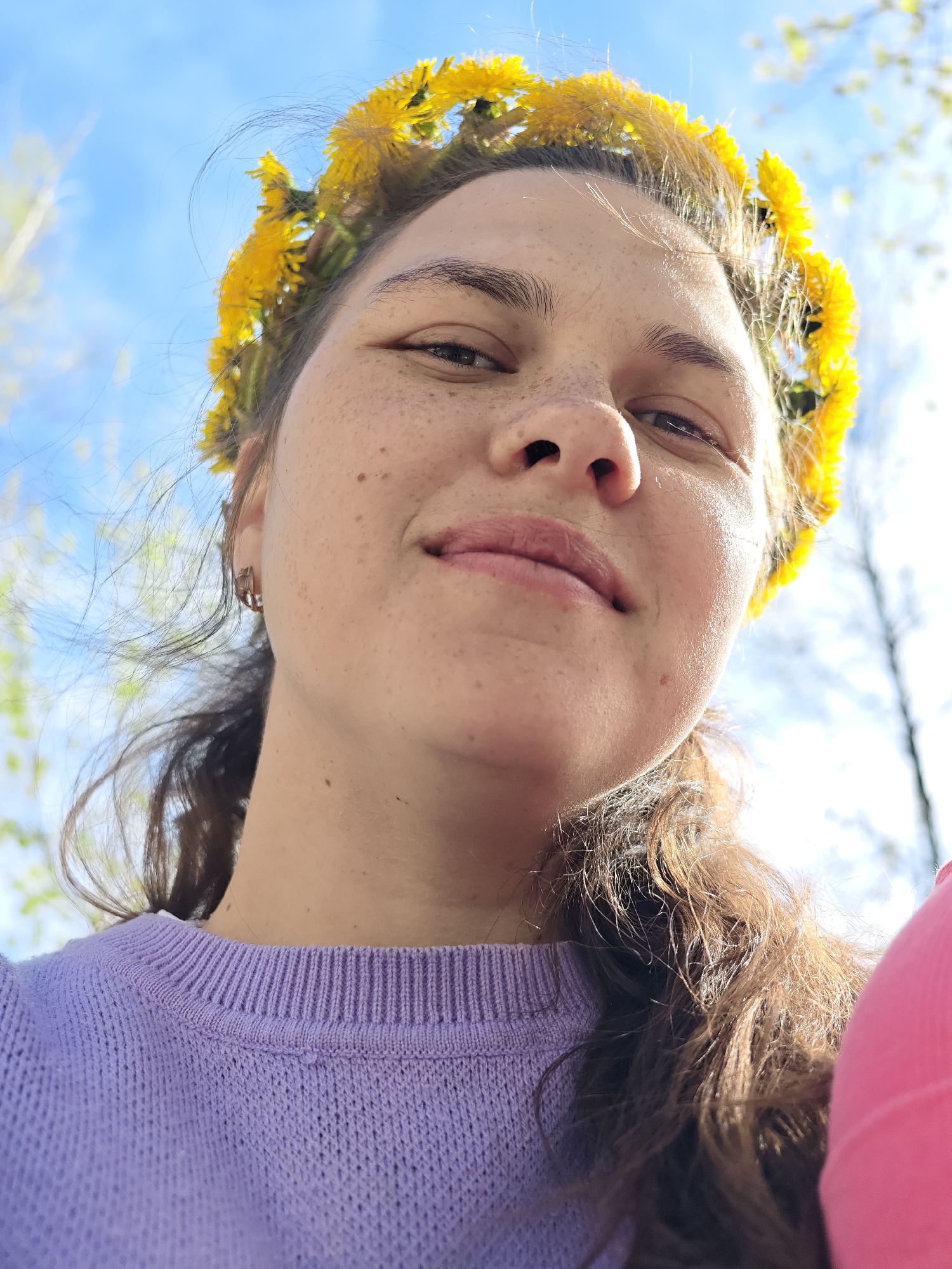 A selfie shows a fair-skinned person with freckles, wearing a crown of yellow dandelions. They are wearing a light purple sweater. Part of a pink garment is visible to the right. The background is a bright, partly cloudy blue sky and some blurry trees. The location is likely in Chaikovsky, Russia.
