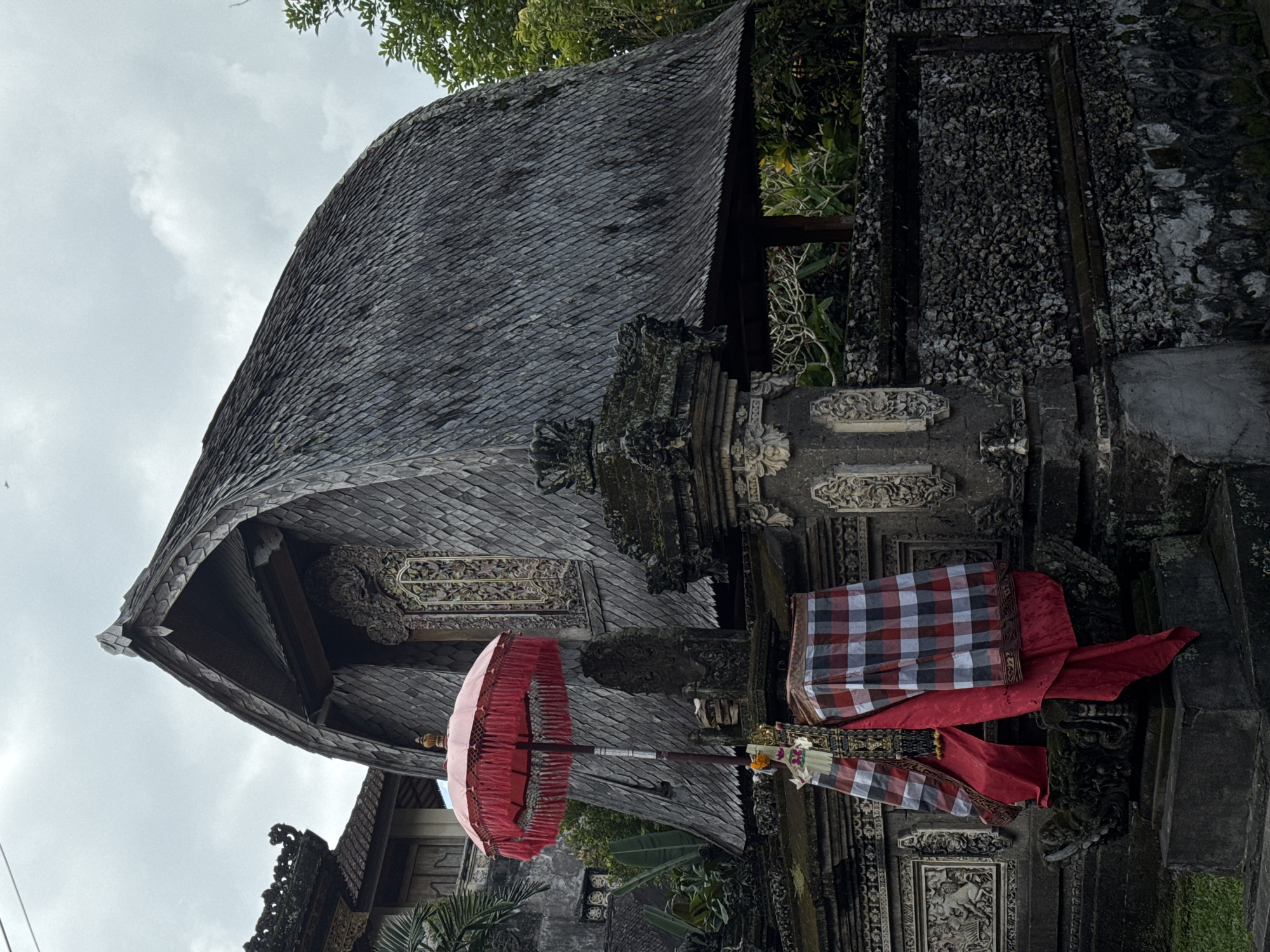 The image shows a traditional Balinese structure with a distinctive thatched roof, located in the Ubud District of Indonesia. The building is made of dark wood and features intricate carvings around a window. In front of the building, a red umbrella with decorative fringe stands on a pole. The structure is surrounded by stone walls with detailed relief carvings and a facade covered in small stones. Red and white checkered fabric is draped over a stone altar adorned with a red cloth. Lush green vegetation is visible in the background, and the sky is overcast. The scene depicts an architectural element of Balinese culture, likely part of a temple or traditional compound.