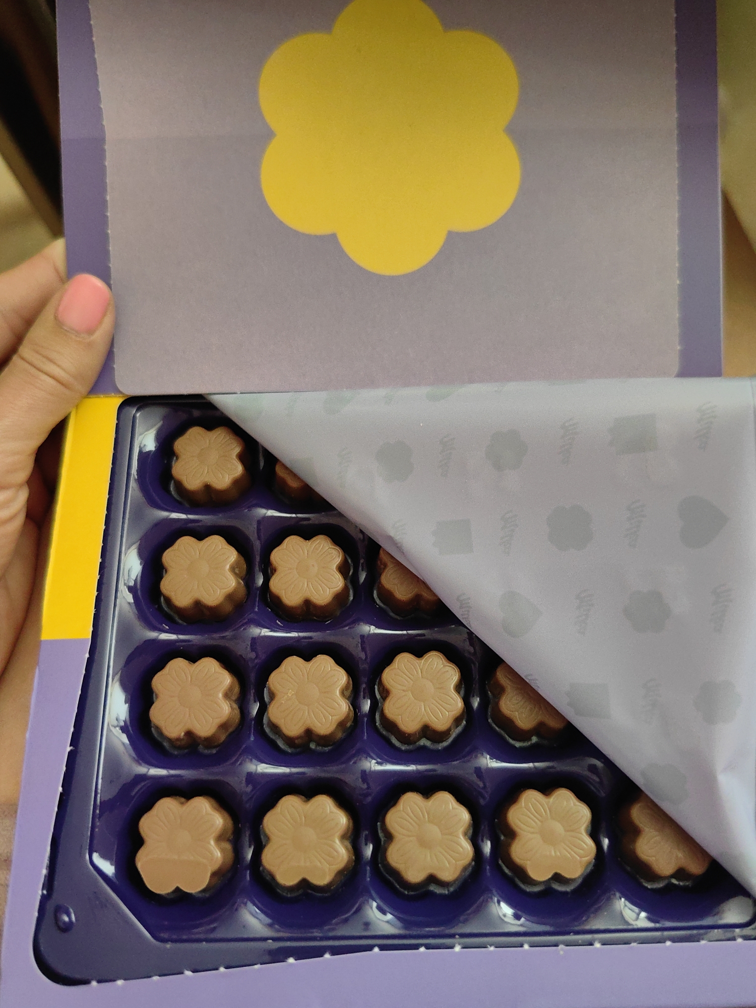 A person's hand with a pink fingernail is holding open a purple box of chocolates. Inside the box, there are multiple rows of flower-shaped milk chocolates. A piece of purple paper with a yellow flower design is folded back, revealing the chocolates. The box is set against a blurred background that suggests an indoor environment. The image does not provide any information about the location in Myadzyel, Belarus.