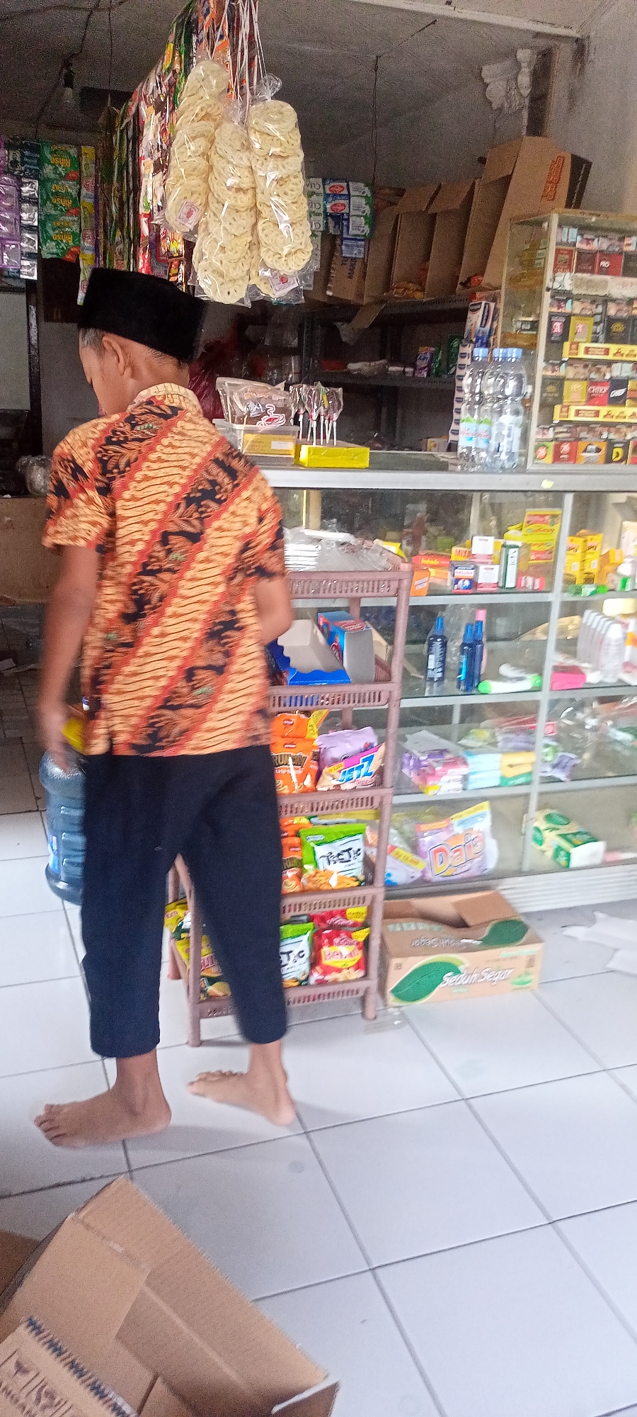 In Gempolklutuk, Indonesia, a person stands in a small convenience store. They are wearing a batik shirt and black pants, with a black peci cap on their head. The person's back is to the camera, and they are looking towards shelves stocked with various goods. The shelves are filled with packaged snacks, drinks, and other items commonly found in a local warung or mini-mart. In the foreground, a large blue water jug is visible. The floor is tiled, and several cardboard boxes are present, one in the foreground containing products. The store's interior appears dimly lit, with goods displayed on shelves and hanging from the ceiling.