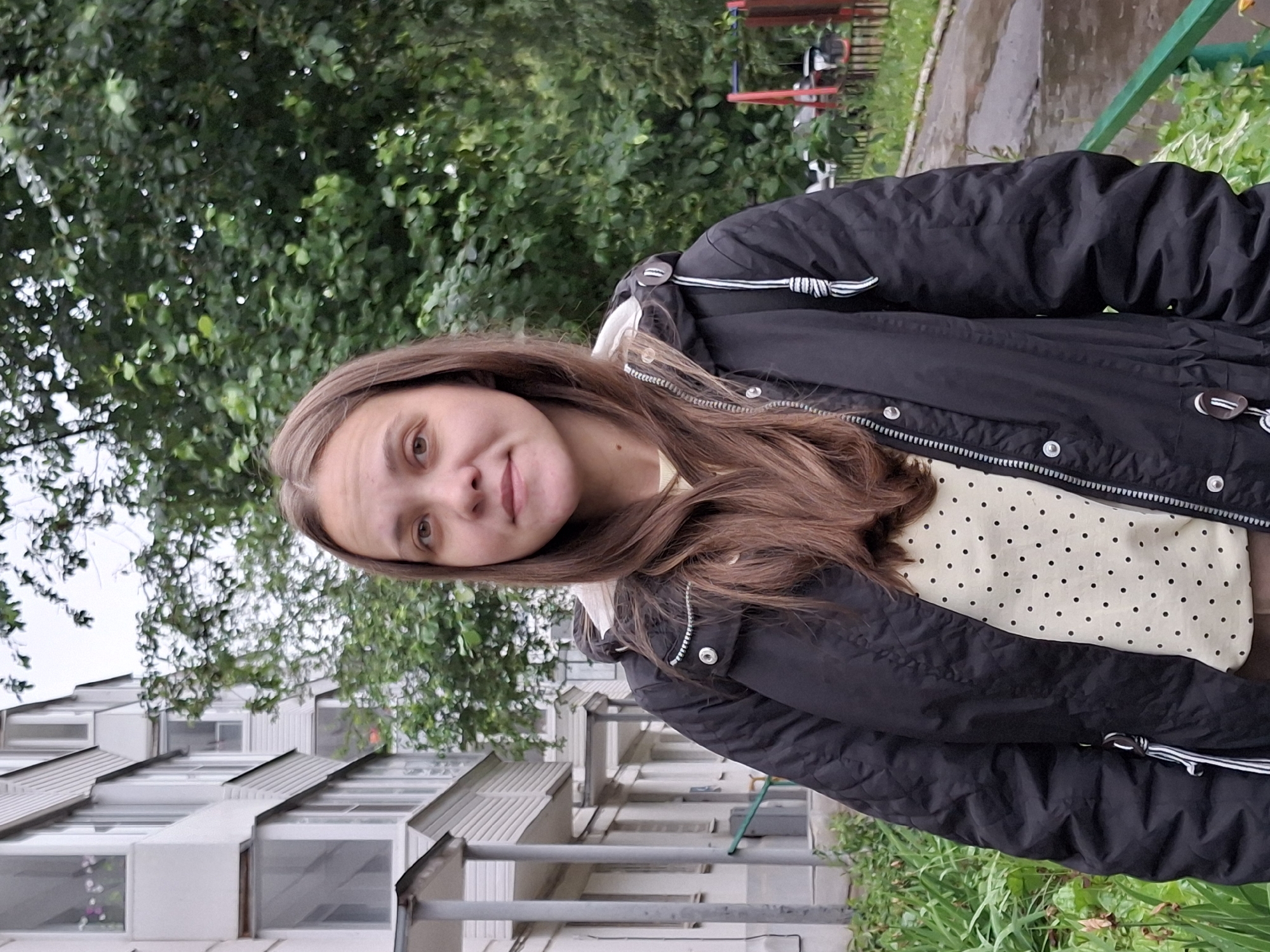 The media file shows a young woman posing for a selfie outdoors. She is wearing a black puffer jacket with white trim and a cream-colored shirt with a black polka dot pattern. Her long, brown hair is visible, falling over her shoulders. The background features a building with a distinctive sloped roofline and multiple windows, suggesting an apartment complex. Lush green trees and foliage are also present, with some grass and what appears to be a wet, paved area, possibly a sidewalk or courtyard. A red and white playground structure can be seen in the upper right portion of the image. The overall lighting suggests it might be an overcast day, as there are no strong shadows. The woman's expression is neutral, with a slight smile. The image is oriented vertically, with the top of her head towards the bottom. The location context is Chaikovsky, Russia. There is no visible text in the media file.