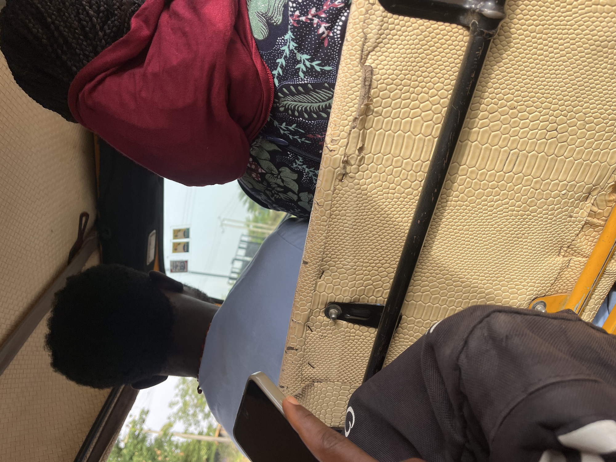 The image, seemingly taken from inside a vehicle in Galadimawa Village, Nigeria, features two individuals and various objects. A person with braided hair wears a maroon top over a floral-patterned garment. Another individual with short, dark hair is seated in front, wearing a light blue shirt and holding a mobile phone. The vehicle's interior includes a faux leather patterned roof, a black metal frame, and a dark gray bag with a white logo. Outside, trees and a building are visible in the background.
