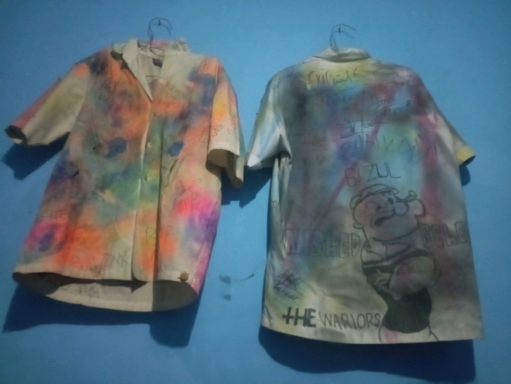 Two shirts, one with a tie-dye effect in various colors and the other with a tie-dye effect and a drawing of Popeye, are hanging on a blue wall. The shirts are on hangers. The image was taken in Senangsari, Indonesia.