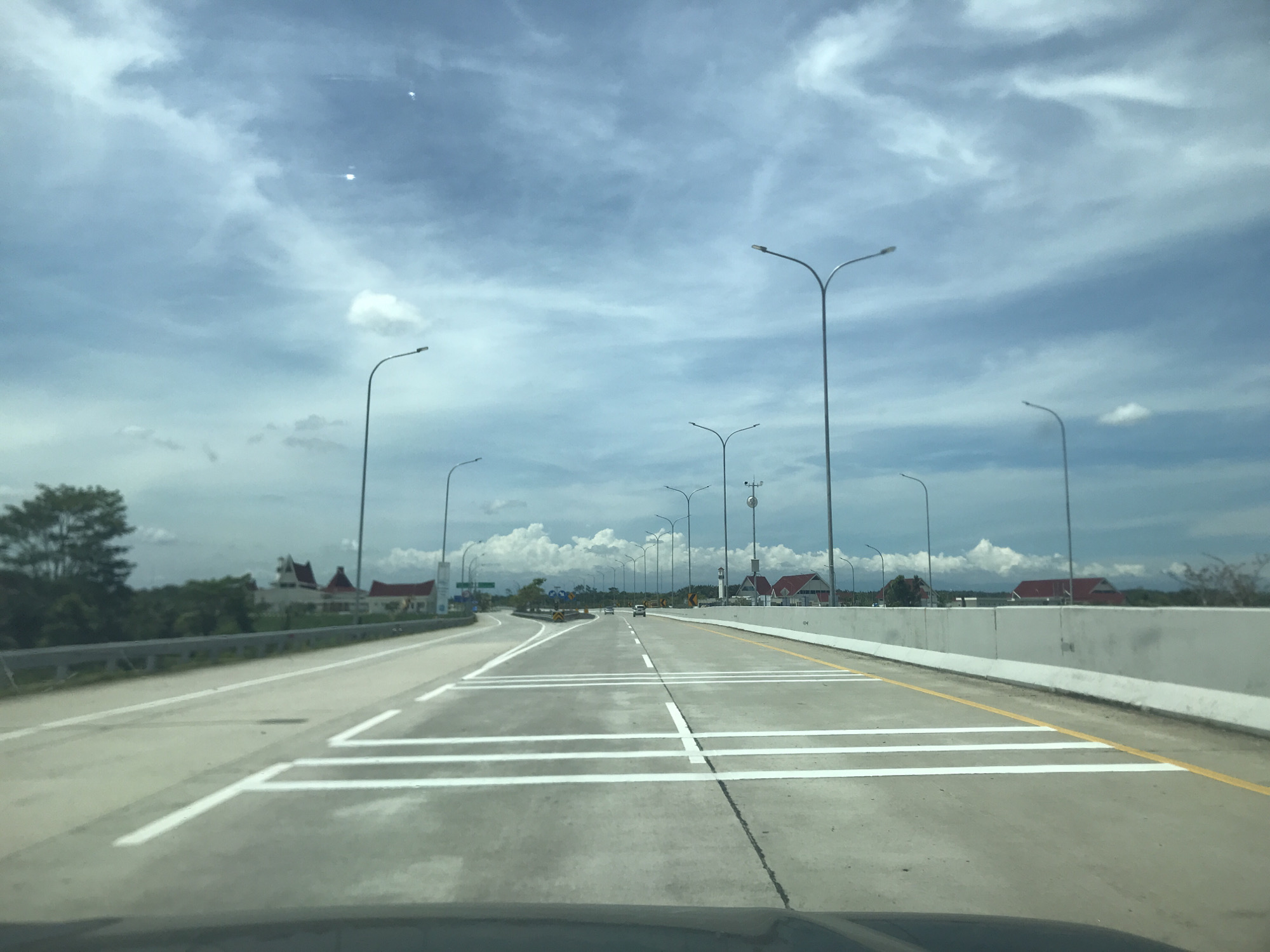 The image shows a view from inside a vehicle driving on a highway in Gohor Lama, Indonesia. The road is gray concrete with white lane markings and a yellow shoulder line. There are several tall streetlights lining both sides of the highway. Buildings with red roofs are visible in the distance. The sky is blue with scattered white clouds.