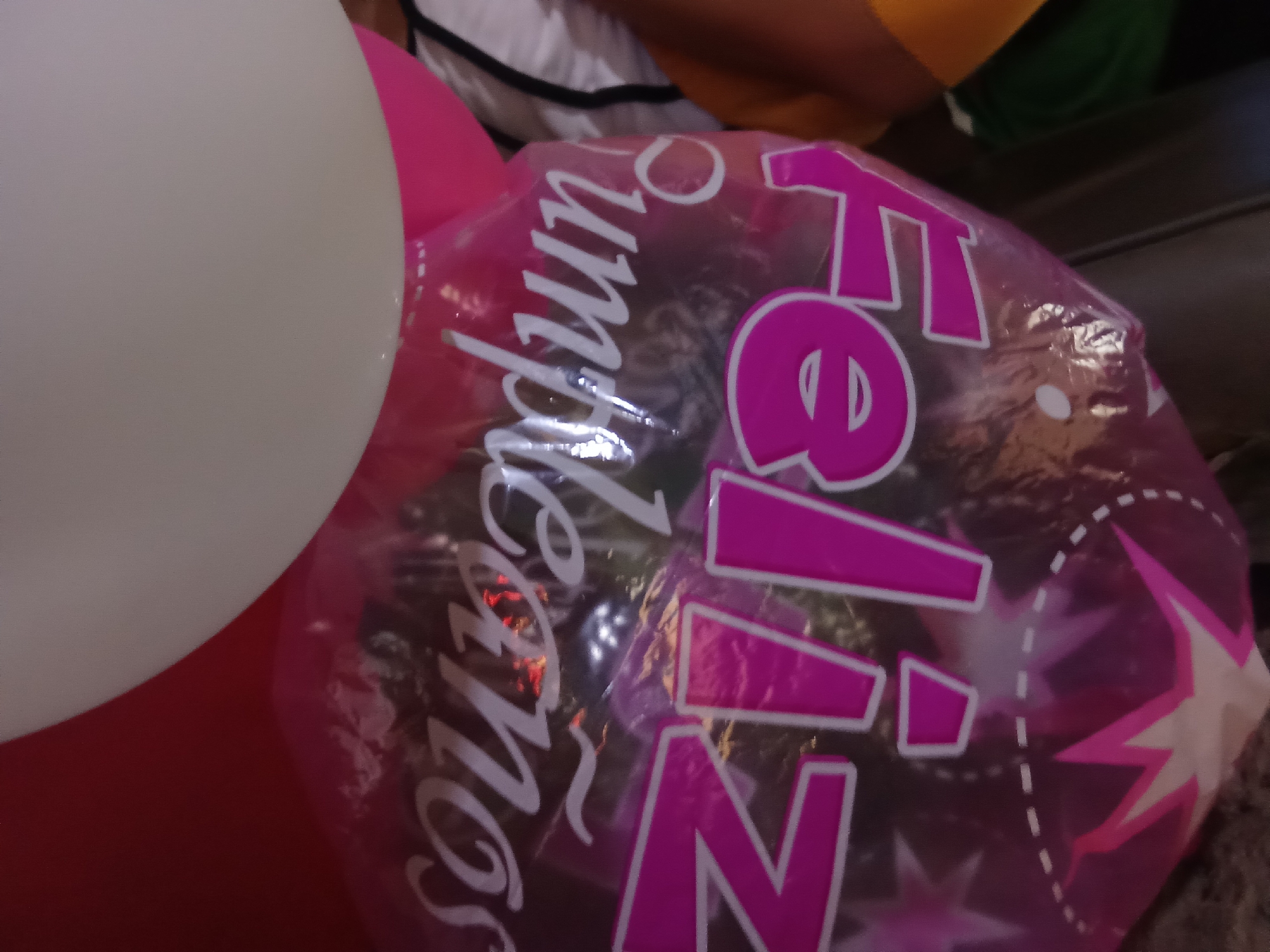 The image shows two partially visible balloons. One is white and the other is a translucent pink with the words "Feliz Cumpleaños" printed on it in pink and white. The word "Feliz" is prominently displayed, and the phrase is accompanied by pink stars. The background is blurred, but it appears to be an indoor setting. The location is Juan Griego, Venezuela.