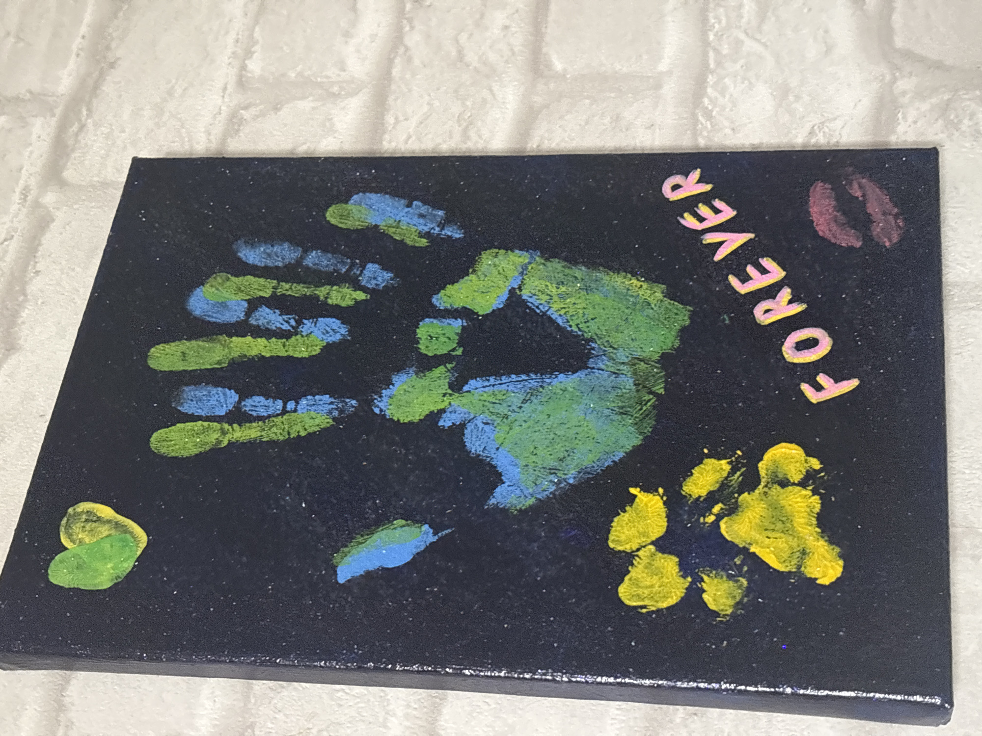 The image shows a black canvas with various painted elements. In the upper left corner, there is a heart shape painted in green and yellow. Dominating the center is a large handprint with fingers pointing upwards, rendered in blue and green paint. Below the handprint, on the left side, are two yellow paw prints. Towards the bottom right of the canvas, the word "FOREVER" is written in pink and yellow letters, and below that, there is a red lipstick kiss print. The canvas is positioned against a white brick wall. No information about the location in the city of Omsk, Russia, is present in the image.