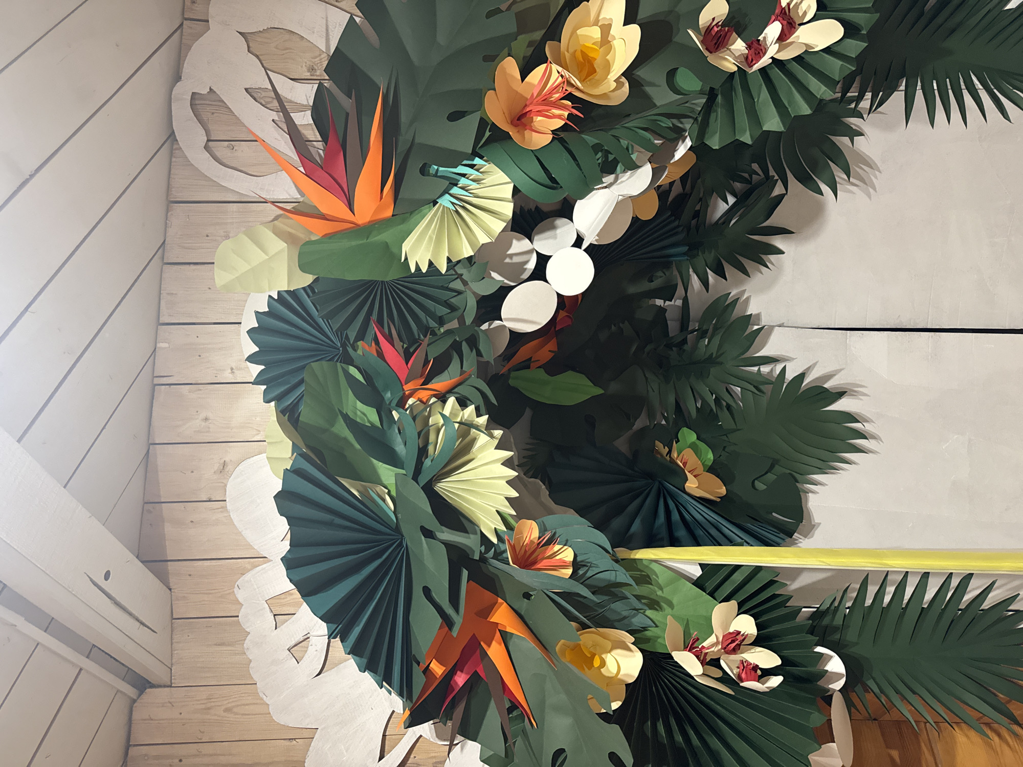 This media file features a vibrant, handcrafted wall decoration composed entirely of paper tropical leaves and flowers. The dense display is arranged in an arch-like or circular pattern, creating a lush, jungle-like aesthetic.
**Subjects:** The primary subjects are numerous paper cutouts representing various botanical elements. These include large, dark green monstera-like leaves, intricate pleated fan leaves in dark green and lighter teal, and slender palm fronds. Interspersed among the foliage are brightly colored paper flowers: vivid orange and red bird of paradise blooms, multi-layered yellow and cream flowers with ruffled centers, and smaller yellow/cream blossoms featuring red stamens. White circular and oval cutouts are also visible, adding a contrasting element.
**Scene/Setting:** The decorations are mounted against a background of light-colored wooden planks, which appear to be part of a wall or a temporary structure. On the left and bottom left, a white, intricately cut wooden or paper overlay with an abstract floral or swirling pattern is visible beneath the main decorations. The setting appears to be indoors, likely an event space, restaurant, or home in Moscow, Russia, given the provided context.
**Activity or event taking place:** There is no specific activity or event depicted, but the elaborate, festive decorations suggest they are part of a themed celebration, a party backdrop, a photo booth, or a decorative display.
**Notable details:** The handcrafted nature of the decorations is prominent, with visible folds, layers, and distinct cut edges. The color palette is rich and tropical, dominated by various shades of green, bright oranges, reds, and yellows, with touches of white. The lighting suggests an indoor environment, possibly with artificial light sources creating soft shadows that enhance the three-dimensional quality of the paper art. No people are visible in the image.
**Visible text:** There is no visible text in the image.