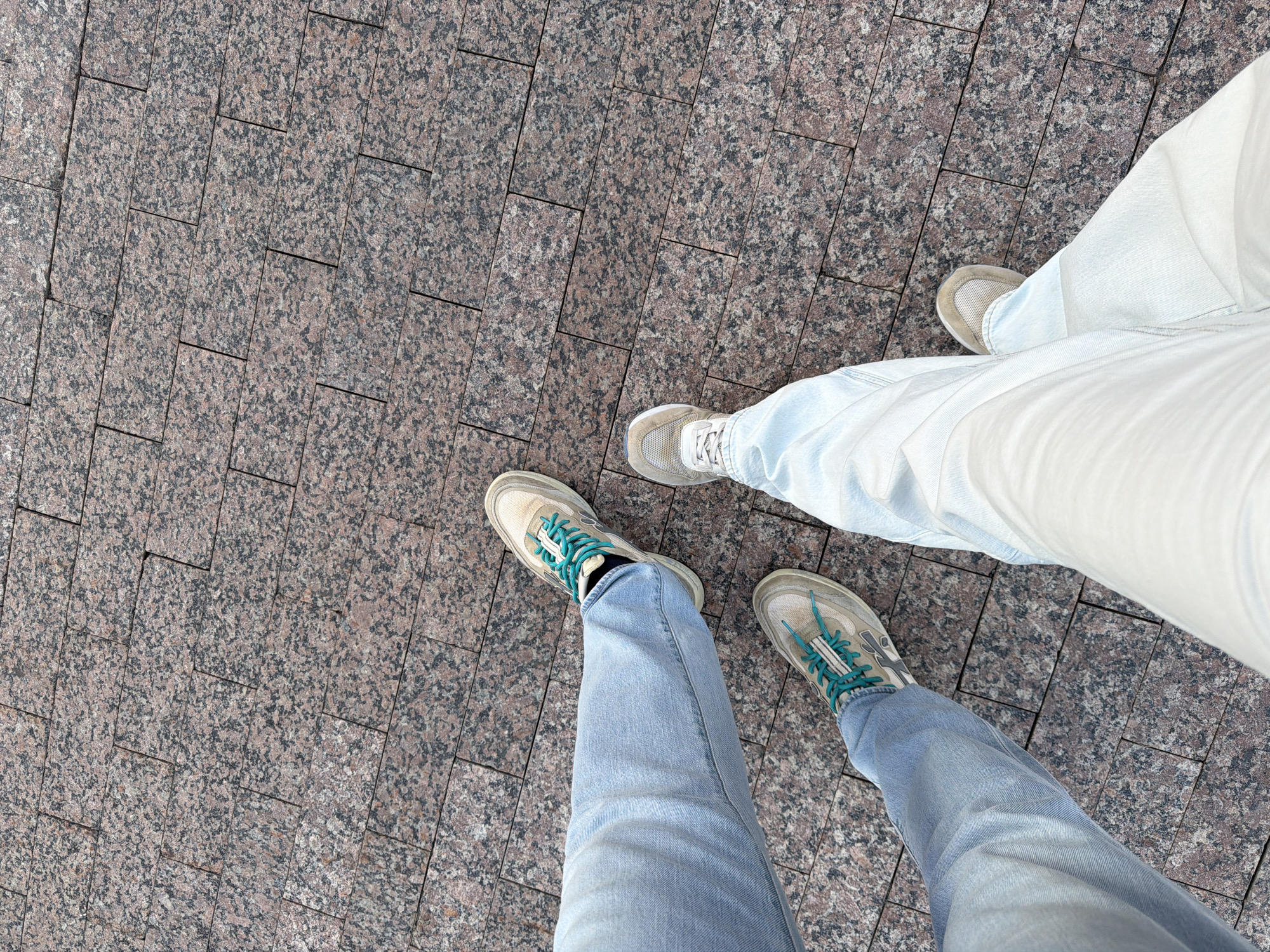 Two pairs of legs, wearing light-wash denim jeans and sneakers, are standing on a sidewalk paved with rectangular granite tiles. The sneakers are low-top athletic shoes, one pair with bright teal laces, and the other with white laces. The granite tiles are speckled with pink, black, and gray. The perspective is a top-down view, focusing on the lower legs and feet. The setting appears to be an urban outdoor environment, consistent with a city street or plaza in Moscow, Russia. The image captures a moment of stillness, with no visible interaction between the individuals.