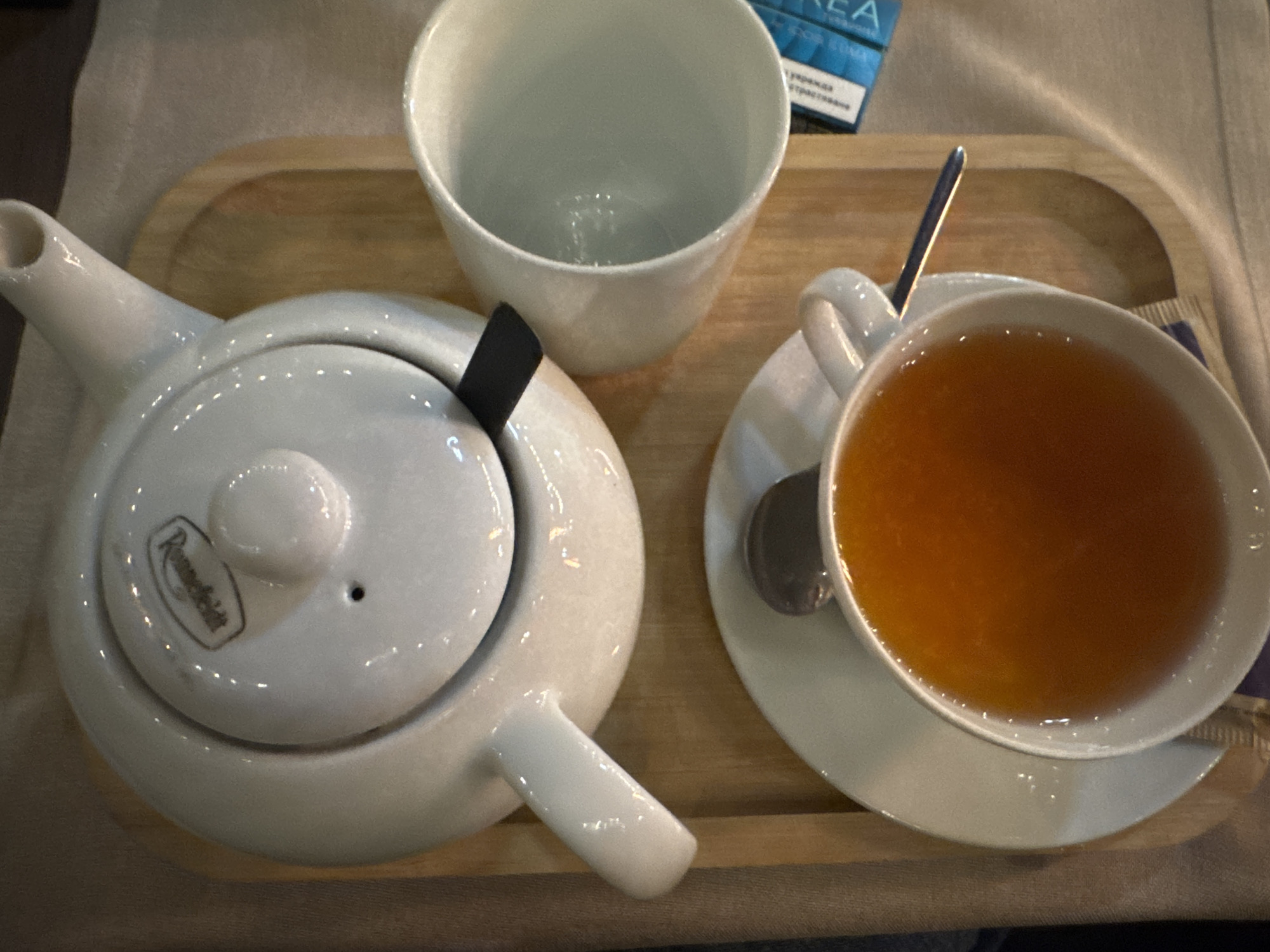 An overhead view displays a tea service situated on a light brown wooden rectangular tray with rounded corners. A white ceramic teapot, marked with the "Ronnefeldt" brand name on its lid, is positioned at the top of the tray, with a black element visible beneath its lid. To the immediate right of the teapot, an empty white ceramic cup stands vertically. At the bottom of the tray, a white ceramic teacup filled with an amber-colored liquid is placed on a matching white saucer, with a silver spoon resting inside the cup. A blue and white rectangular package is partially visible to the right of the empty cup, and a dark blue wrapper is present beneath the saucer of the filled teacup. The tray rests on a light-colored fabric surface. The scene is located in the city of Unknown, Unknown.