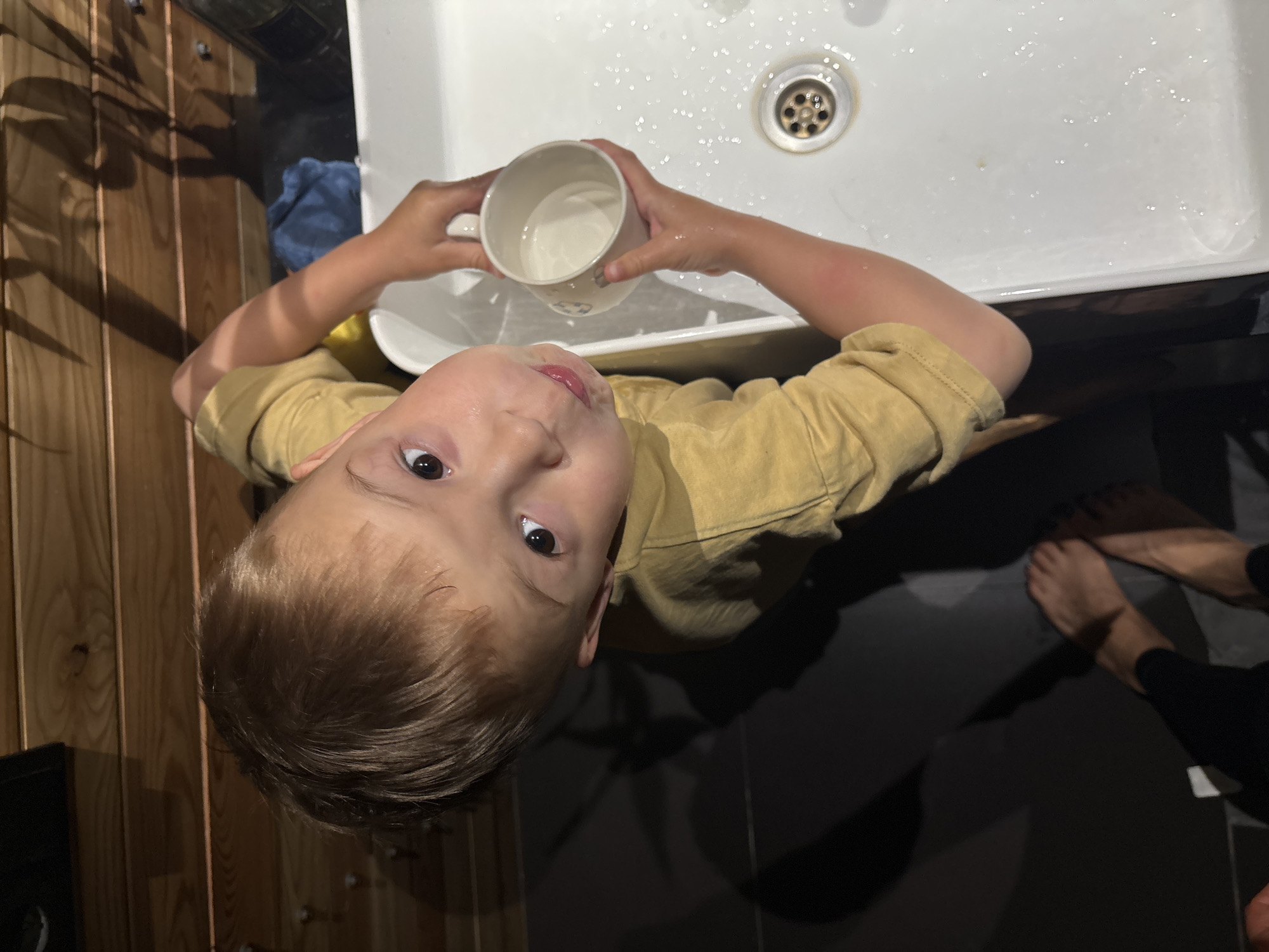 A young boy is positioned above a white sink, holding a white mug filled with liquid. He is wearing a light brown shirt and looking up at the camera. The sink has a drain in the center and some water droplets. Wooden panels are visible on the left side, and part of a person's feet are on the right side. The setting is a bathroom. It is unknown whether this image was taken in the city of Chaikovsky, Russia.