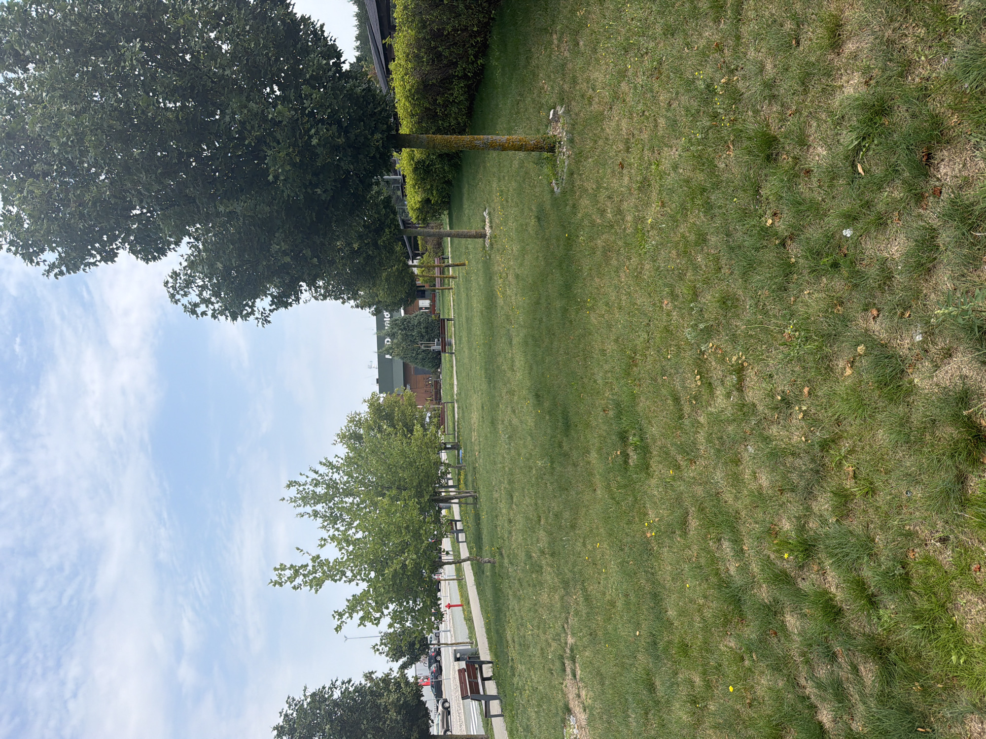 The image depicts an outdoor scene with a grassy park area in the foreground, featuring several trees, benches, and a paved pathway. In the background, a building with a McDonald's sign is visible, suggesting the location is near a McDonald's restaurant. The sky is partly cloudy. The environment appears to be a public space, possibly a rest stop or a recreational area adjacent to a commercial establishment. There are no people visible in the image. The overall scene suggests a pleasant, albeit not bustling, day.