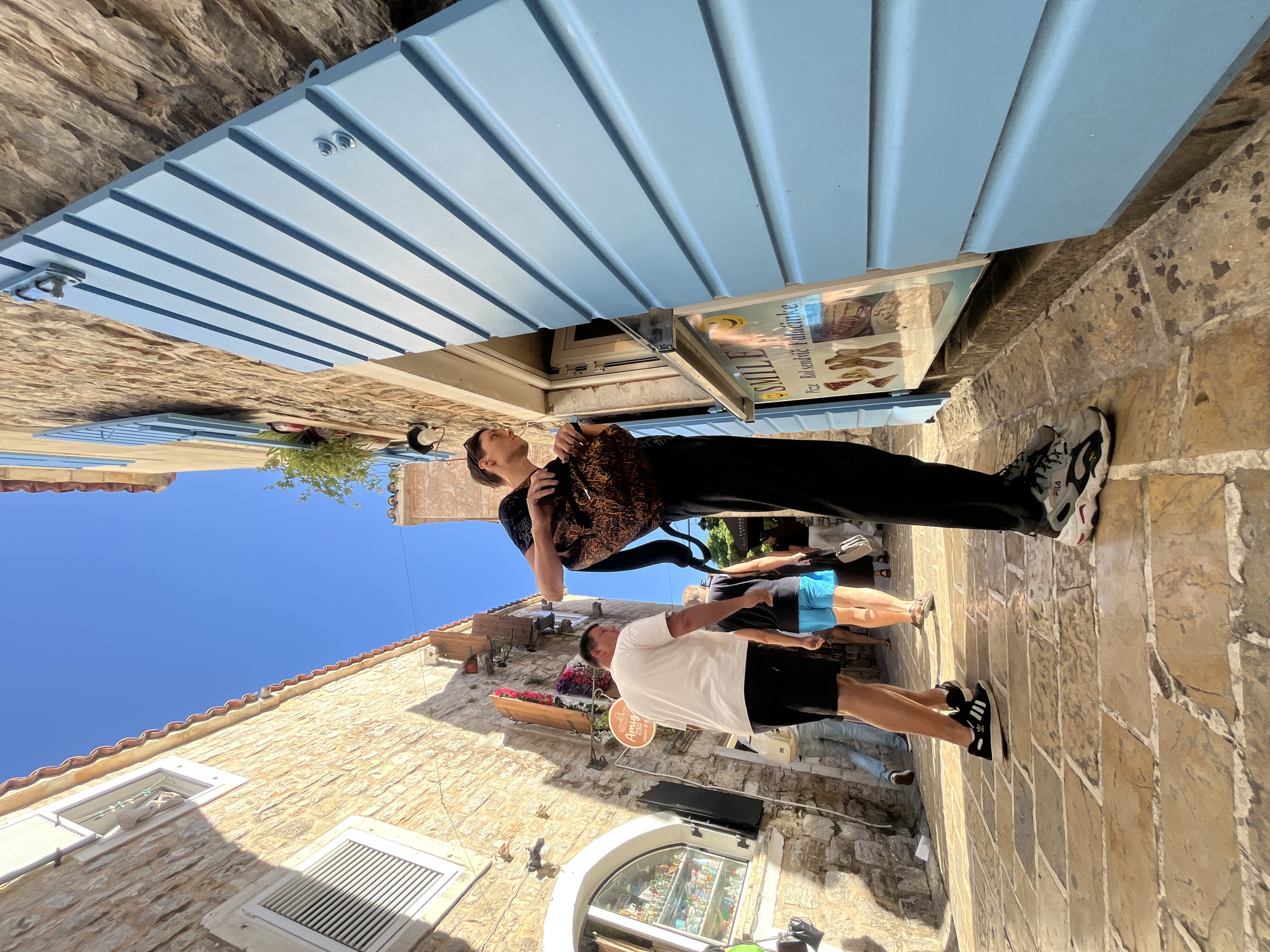 This image captures a vibrant, sun-drenched stone alleyway in Budva, Montenegro, viewed from a low angle looking upwards towards the clear blue sky. The scene is illuminated by bright daylight, casting distinct shadows and highlighting the texture of the old stone buildings.
In the foreground, a person dressed in a dark, patterned long-sleeved shirt and black trousers, carrying a black backpack, stands facing towards the left, looking intently upwards. To their right and slightly behind, another individual in a white t-shirt, black shorts, and dark sneakers walks or stands on the cobblestone path, facing away from the camera. Other people are partially visible further down the alleyway, suggesting foot traffic.
The alley is lined with traditional buildings constructed from light brown, textured stone. A prominent feature is a light blue, slatted wooden awning or shutter structure extending from a building overhead, showing visible bolts and hinges. Below this structure, a sign is attached, displaying the word "SMILE" and partially legible text "Rule & sundrie valami ke." Further down the street, a circular sign with "Amay" and "OTBOS" is visible. Windows with white grates and a decorative flower box add to the charm of the architecture. The ground is paved with varied light-colored cobblestones, reflecting the strong sunlight.
The clear, deep blue sky is visible above the buildings, indicating a bright and sunny day. The scene captures the lively atmosphere of an old town street under warm weather.
