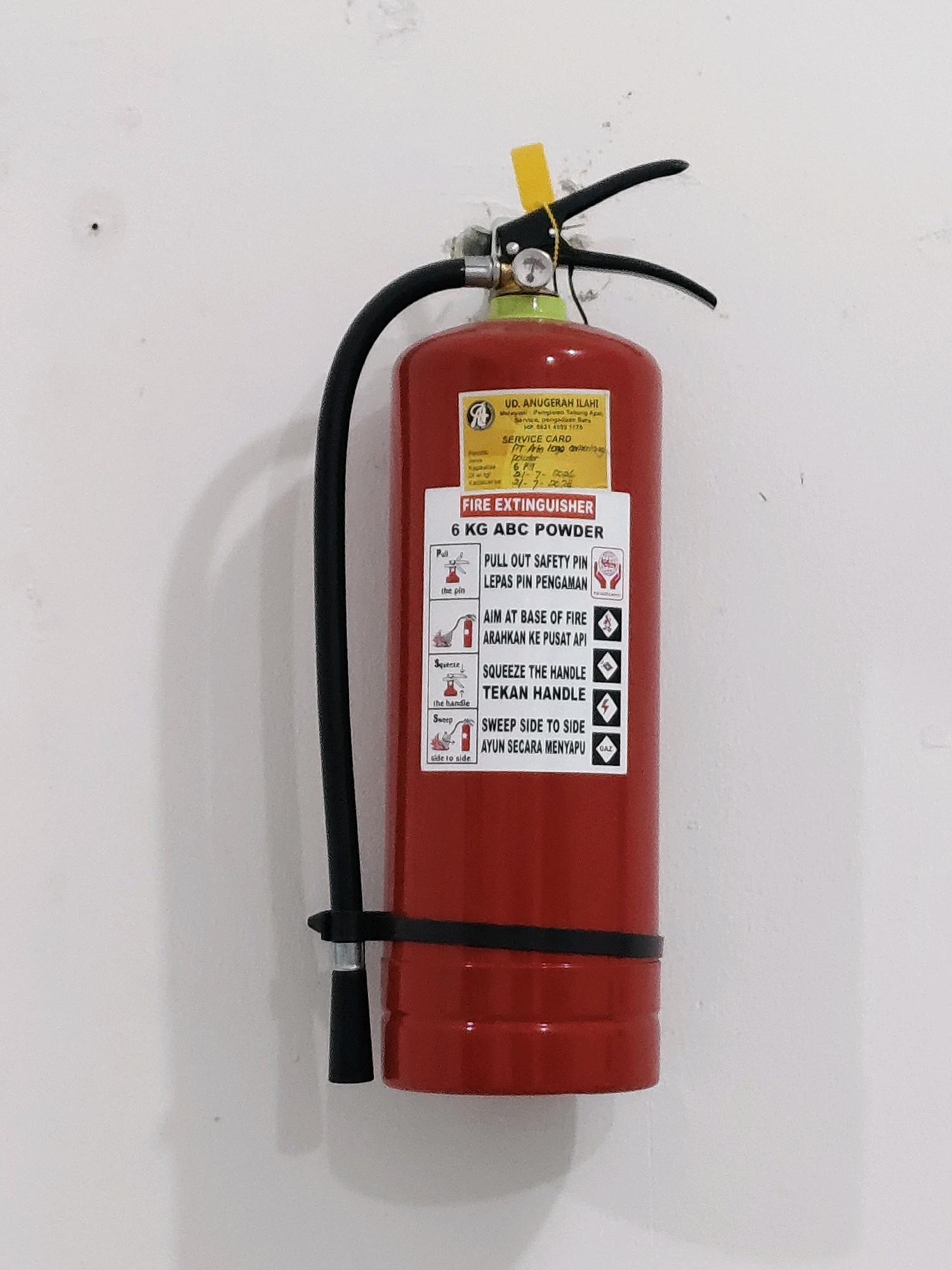 A red, 6 kg ABC powder fire extinguisher is mounted on a white wall. The extinguisher has a black hose and handle assembly. A yellow tag is attached to the safety pin. A label on the extinguisher provides instructions in English and Malay for its use. The label also includes information about the distributor, UD. ANUGERAH ILAHI, and a service card. The wall appears to be textured and has some minor imperfections. The image was taken indoors. The location of the image is in the city of Probolinggo, Indonesia.