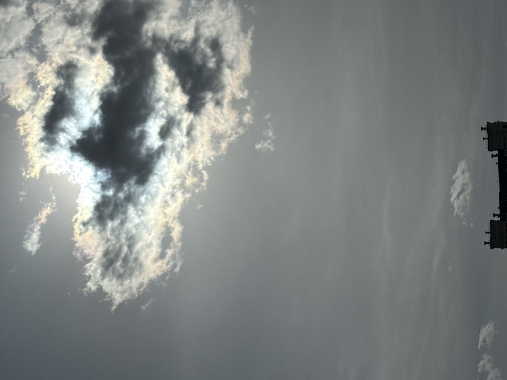 The image displays a dramatic sky scene dominated by thick, dark clouds. The sun, partially obscured by these clouds, shines brightly through an opening on the left side, creating a radiant highlight. The edges of the darker clouds surrounding this bright area exhibit subtle iridescence, with hints of rainbow colors due to light diffraction. The sky around these main cloud formations is a muted, lighter grey, containing some faint, wispy cloud textures.
On the far right side of the frame, a dark, rectangular architectural structure is partially visible. This structure, which appears to be part of a building, a billboard, or a tower, is silhouetted against the lighter sky and features some small, protruding elements along its top edge. The scene depicts a daytime sky with cloudy weather and strong backlighting from the sun. There are no people visible, and no discernible text. The overall impression is one of natural light interacting with atmospheric conditions.