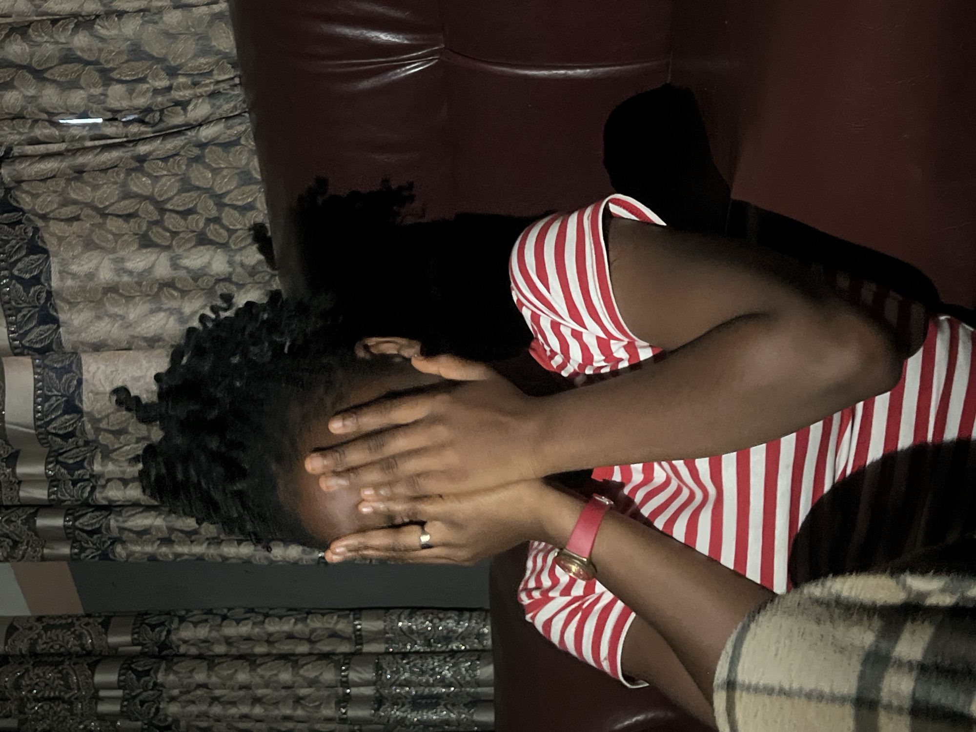 The image shows a person, appearing to be a woman with dark skin and dark, curly hair, sitting on a dark red couch. Her face is covered with her hands, suggesting she is hiding or embarrassed. She is wearing a red and white striped shirt and a watch with a pink band on her left wrist. To her left, a checkered blanket or cloth is visible. In the background, patterned curtains hang. The scene appears to be indoors, likely in a living room. The image is dimly lit, with a strong light source casting shadows. The person's location is described as being in the city of Nkpor, Nigeria.