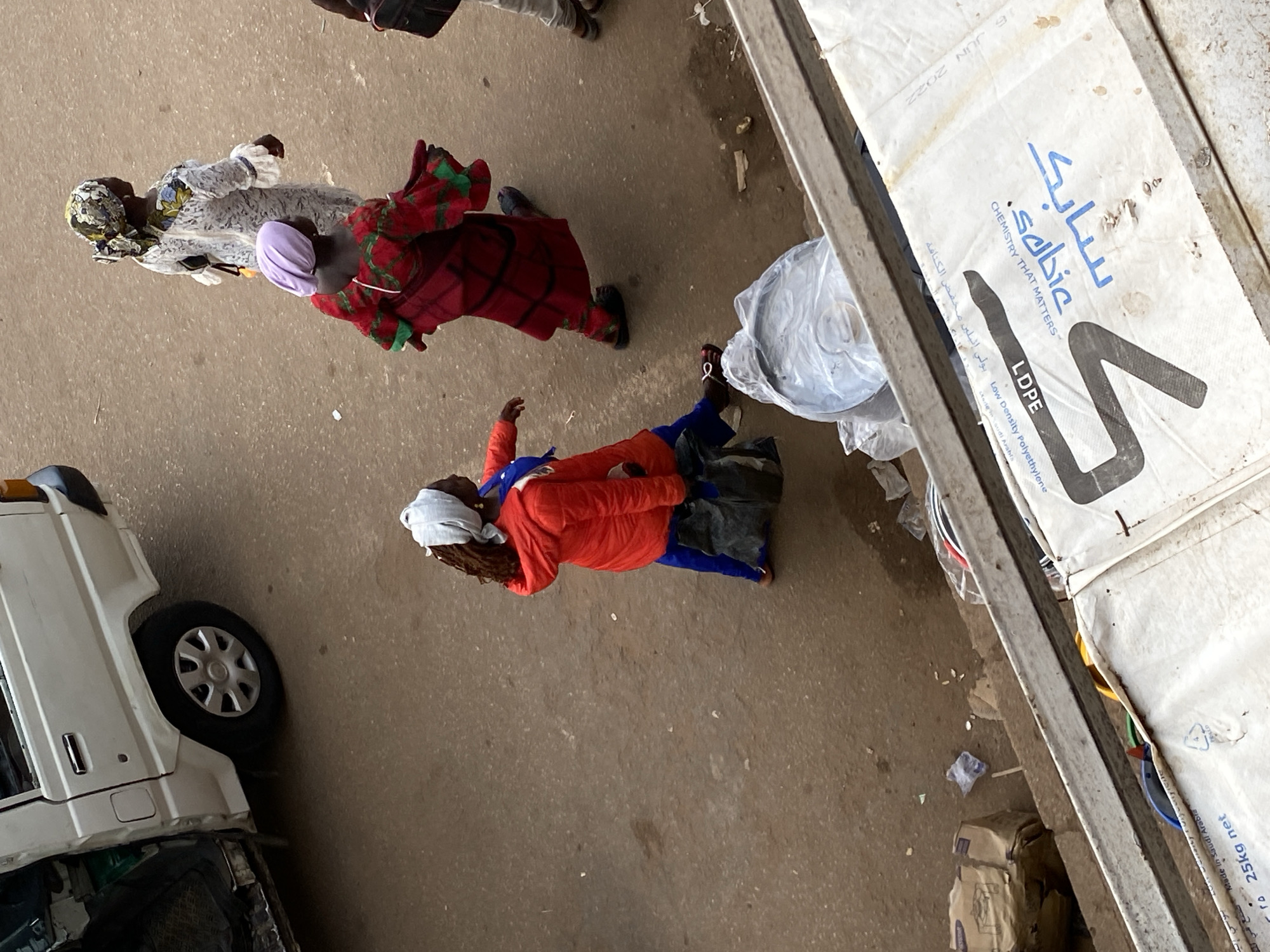 This overhead image depicts a street scene in the city of Bukuru, Nigeria. The primary focus is on pedestrian activity and a stationary vehicle on a dusty, light brown road surface.
Three individuals, all appearing to be female, are walking on the road. In the mid-foreground, a person wears a bright red hooded jacket over blue pants, with a white headwrap covering their braided hair. They are carrying a dark bag in their left hand and wear light-colored sandals. Ahead and to their right, another person wears a purple headwrap and a loose, flowing red garment with green patterns, walking away from the camera in dark sandals. Further ahead and to the right, a third person in a patterned white dress with long sleeves and a multicolored headwrap is also walking away, carrying a dark shoulder bag. The lower legs and feet of a fourth person in light pants and brown footwear are partially visible on the far right edge, walking towards the top of the frame.
In the upper left quadrant, a white minivan or small bus is parked, with its rear passenger door open, revealing a dark interior. The vehicle's rear wheel, fitted with a silver hubcap, is clearly visible.
The ground surface is a mix of compacted earth and possibly degraded asphalt, with scattered small pieces of light-colored debris. Across the bottom third of the image, a metal railing or beam indicates an elevated viewing position. Below this railing and to the right, several large white industrial sacks are visible, one of which displays text in Arabic and English, including "LDPE," "SABIC," "CHEMISTRY THAT MATTERS," "Made in Saudi Arabia," and a "25kg net" weight, along with a date of "16 JUN 2022." Transparent plastic bags containing unidentifiable contents and cardboard boxes are also present below the railing.