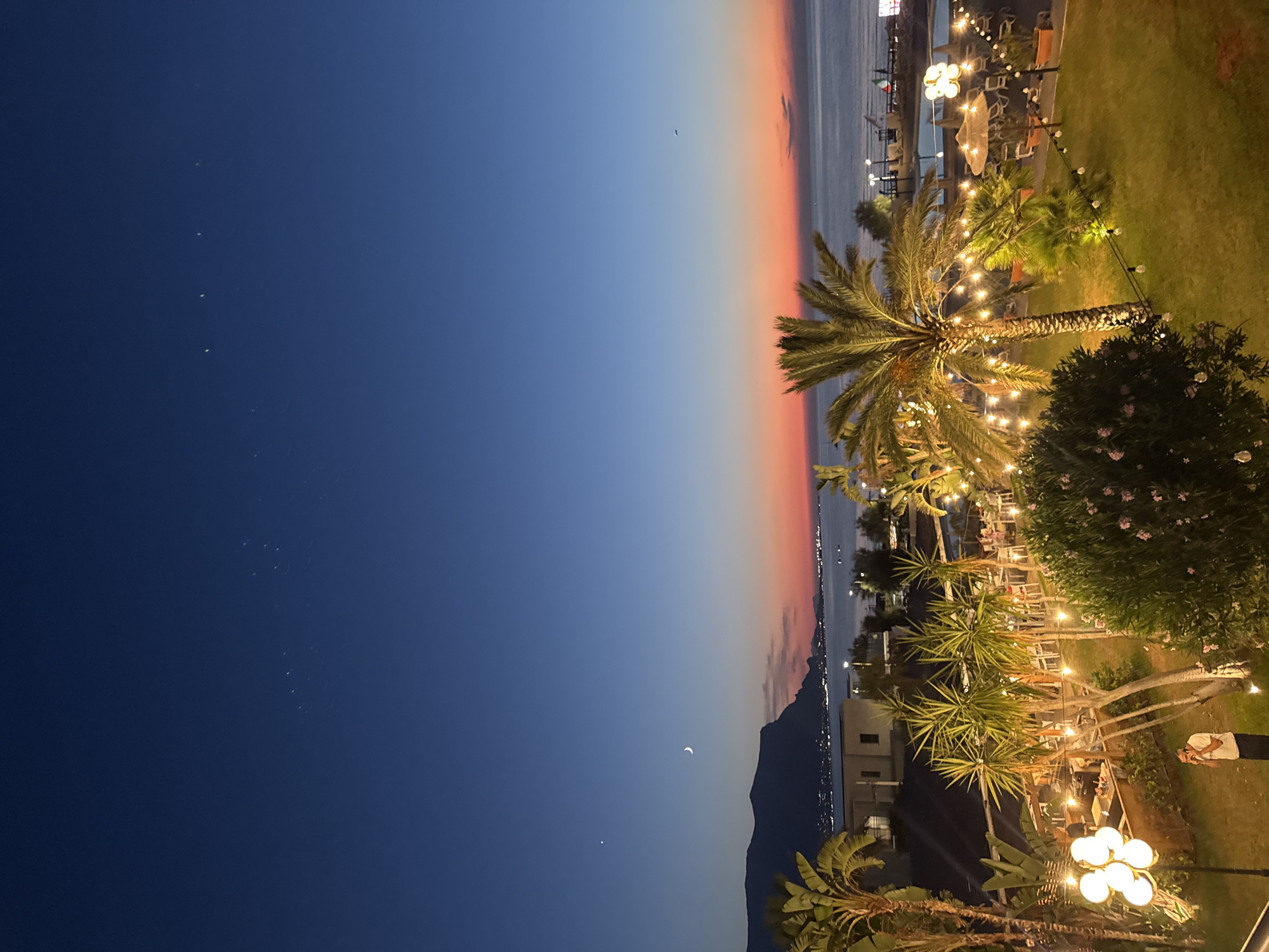 This image depicts a twilight scene on the coast of Isola delle Femmine, Italy. The sky transitions from a deep blue to hues of orange and pink near the horizon, where the sea meets the land. A crescent moon is visible in the upper left quadrant of the sky. In the foreground, a well-lit garden area is populated with palm trees and lush greenery. String lights are draped across the trees and pathways, illuminating the scene. Several tables and chairs are set up, suggesting an outdoor dining or seating area. In the middle ground, buildings line the coast, with lights twinkling from them. A mountain range forms the backdrop on the left side of the image. The sea appears calm, with a few lights visible on the water and along the shore. One person, wearing a white shirt and dark pants, is standing in the lower left corner of the image, appearing to be on their phone.