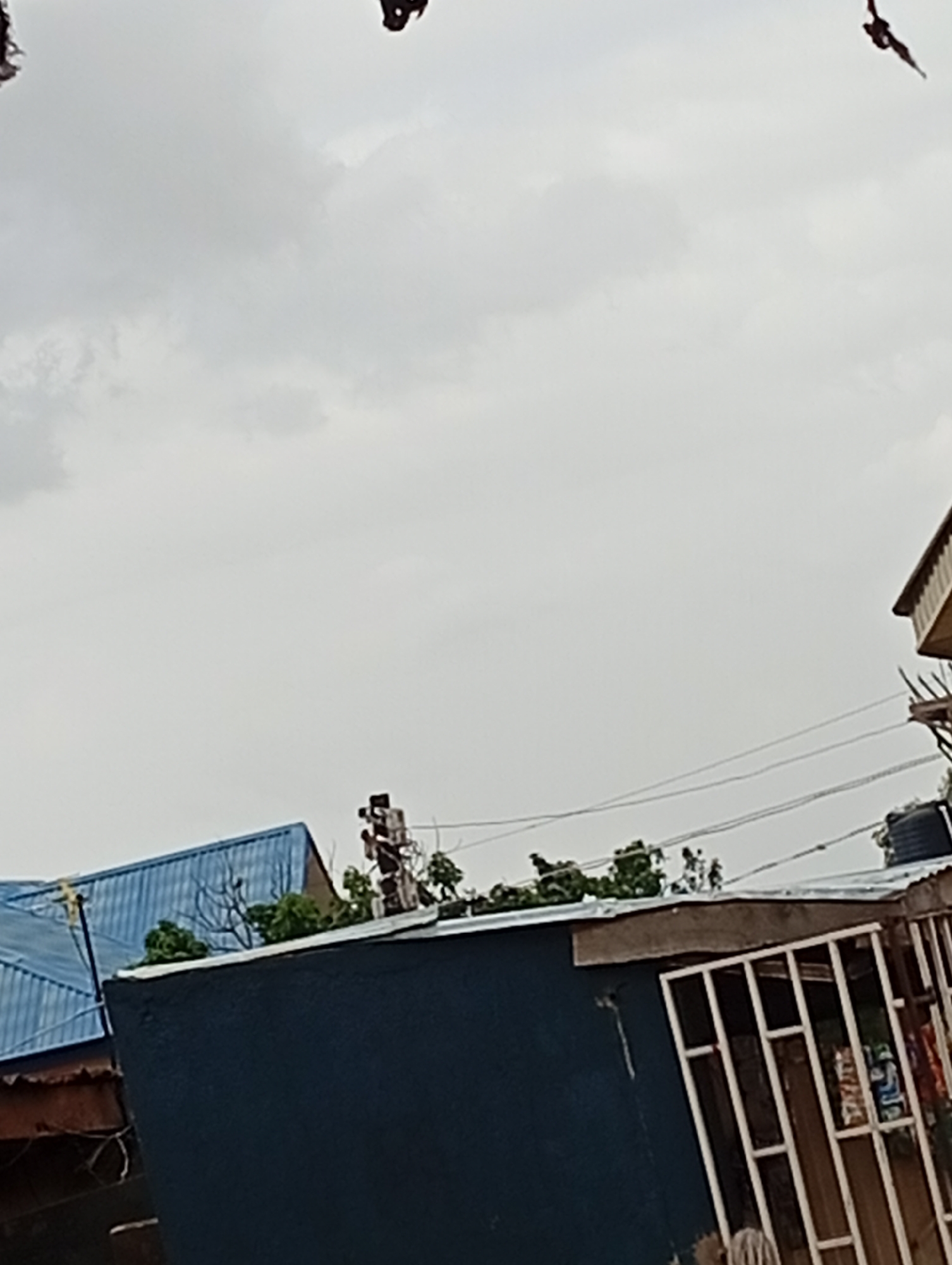 The image shows an overcast sky above a collection of residential buildings in Gura Top, Nigeria. In the foreground, a dark blue structure with a white, barred window is visible. To the left, a building with a blue corrugated metal roof is partially seen. In the background, a utility pole with electrical wires stands amongst some greenery. The top of another building with a lighter colored roof and wooden trim is also present. In the sky, two dark, silhouetted figures, possibly birds or kites, are visible.