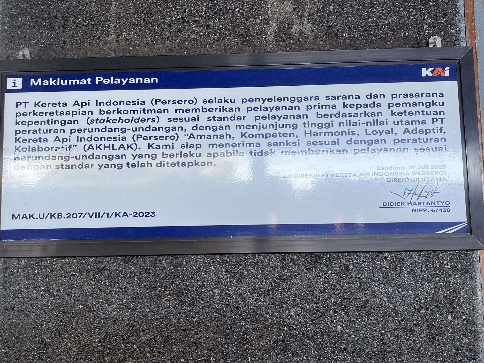 A framed sign is mounted outdoors, likely on a wall or fence. The sign has a blue header with white text that reads "i Maklumat Pelayanan" on the left and the red and white "KAI" logo on the right. Below the header, white text on a light blue background details the commitment of PT Kereta Api Indonesia (Persero) to providing excellent service according to regulations and principles. The bottom of the sign features a registration number "MAK.U/KB.207/VII/1/KA-2023" on the left and a signature, name "DIDIEK HARTANTYO", and NIPP number on the right, dated "Bandung, 27 Juli 2023". The sign is framed in black and mounted against a textured, dark grey surface that appears to be stone or gravel. The location is identified as Desa Tambakreja, Indonesia.