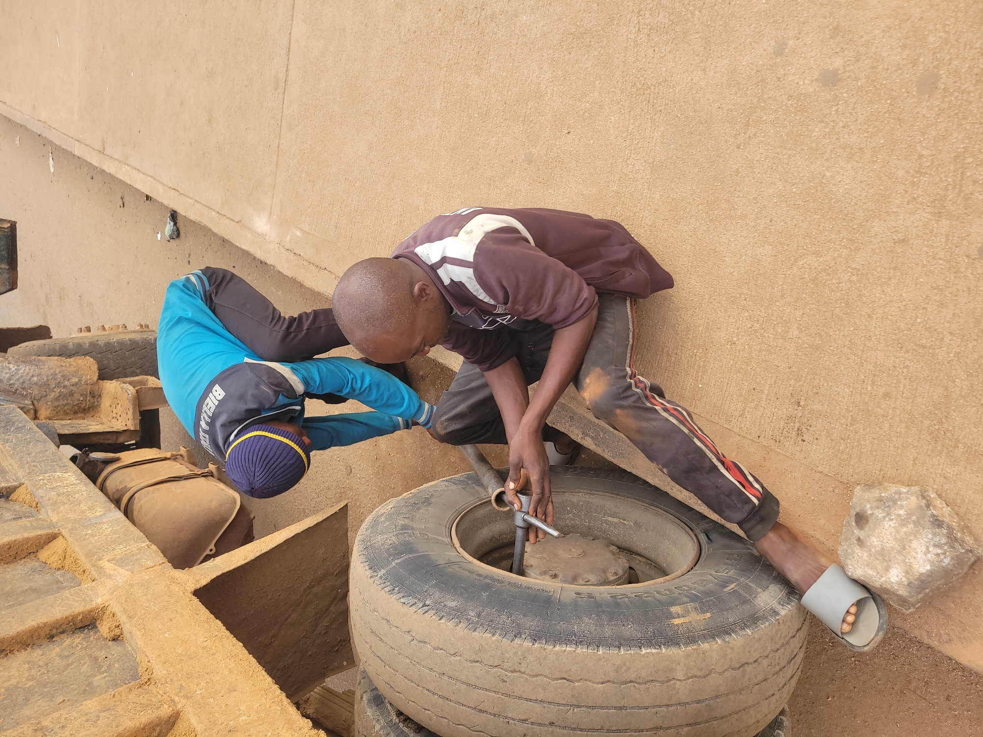 In Gura Top, Nigeria, two individuals are attending to a large truck tire. One person, wearing a blue jacket with a yellow stripe on the collar and a blue beanie, is bent over the truck's chassis. The other person, seated on the ground, is actively working on the tire with a socket wrench, appearing to loosen or tighten lug nuts. The truck is large and appears to be a dump truck, its exterior coated in dirt and sand. The ground is a mix of dirt and a paved surface. Essential details captured include the subjects' clothing, the tool being used, and the context of mechanical work on a large vehicle in an outdoor urban setting.