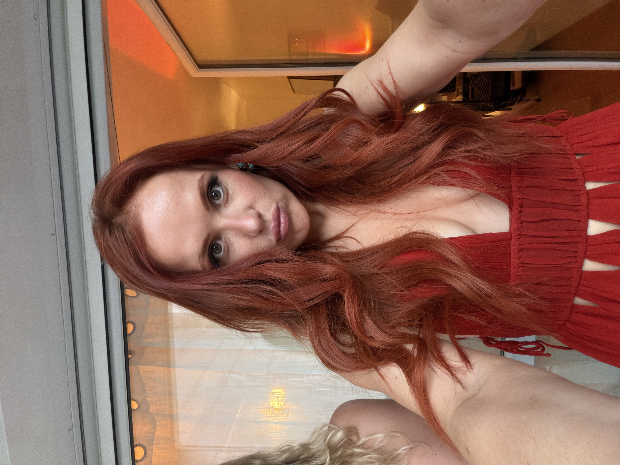 A woman with long red hair poses for a selfie in a red dress. She is inside a room with a window, possibly in Paris, France. The room has a warm, orange light. There is another person with blonde hair partially visible in the bottom of the frame.