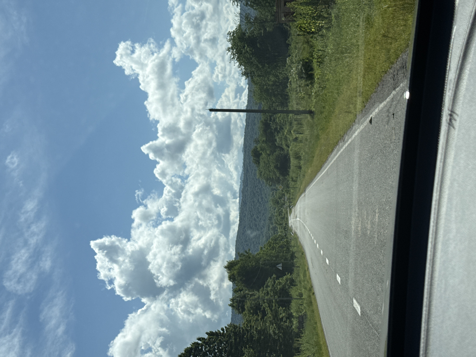 The image depicts a view from inside a vehicle traveling on a paved road in Alagir, Russia. The road is marked with white dashed lines and stretches into the distance towards a tree-covered mountain range. The sky is blue with large, fluffy white clouds. Dense green trees line both sides of the road. On the right side of the road, a tall, dark, slender pole stands prominently in a grassy field, and a section of a brick structure is visible further to the right. The foreground shows the dashboard of the vehicle, partially obscuring the view. The scene suggests a rural or semi-rural setting, possibly a highway or main road leading out of or into Alagir.