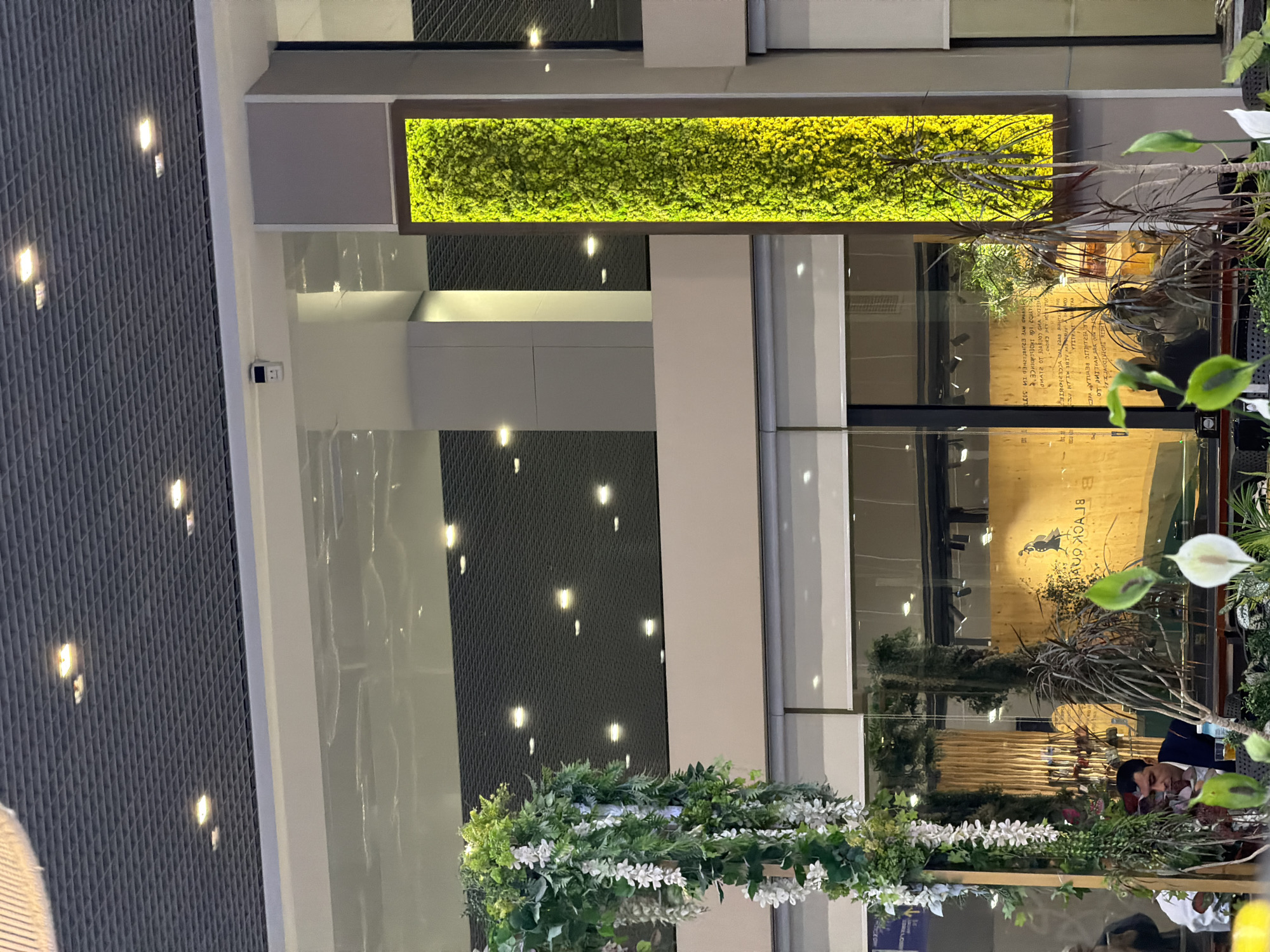 The image depicts the interior of a modern establishment, possibly a cafe or restaurant, featuring an abundance of greenery. A prominent feature is a tall, vertical display of lush green moss or similar plant material, illuminated by a warm yellow light, set against a neutral-colored wall. To the left, an elaborate floral arrangement of white flowers and green foliage cascades downwards, framing the scene. In the mid-ground, through large glass windows, a man in a dark suit is seated at a table, appearing to be engaged in conversation. Behind him, a wooden panel with text is visible, which when reversed appears to read "BLACK GOOSE". The ceiling is a dark, perforated grid with numerous square lights. Reflections on the upper glass surfaces suggest a large, open space, possibly a mall or airport, with more lighting and structural elements visible. The overall ambiance is one of contemporary design with a strong emphasis on natural elements. This scene is located in the city of Tashkent, Uzbekistan, likely within the HUMO Arena complex, known for its modern architecture and event spaces.