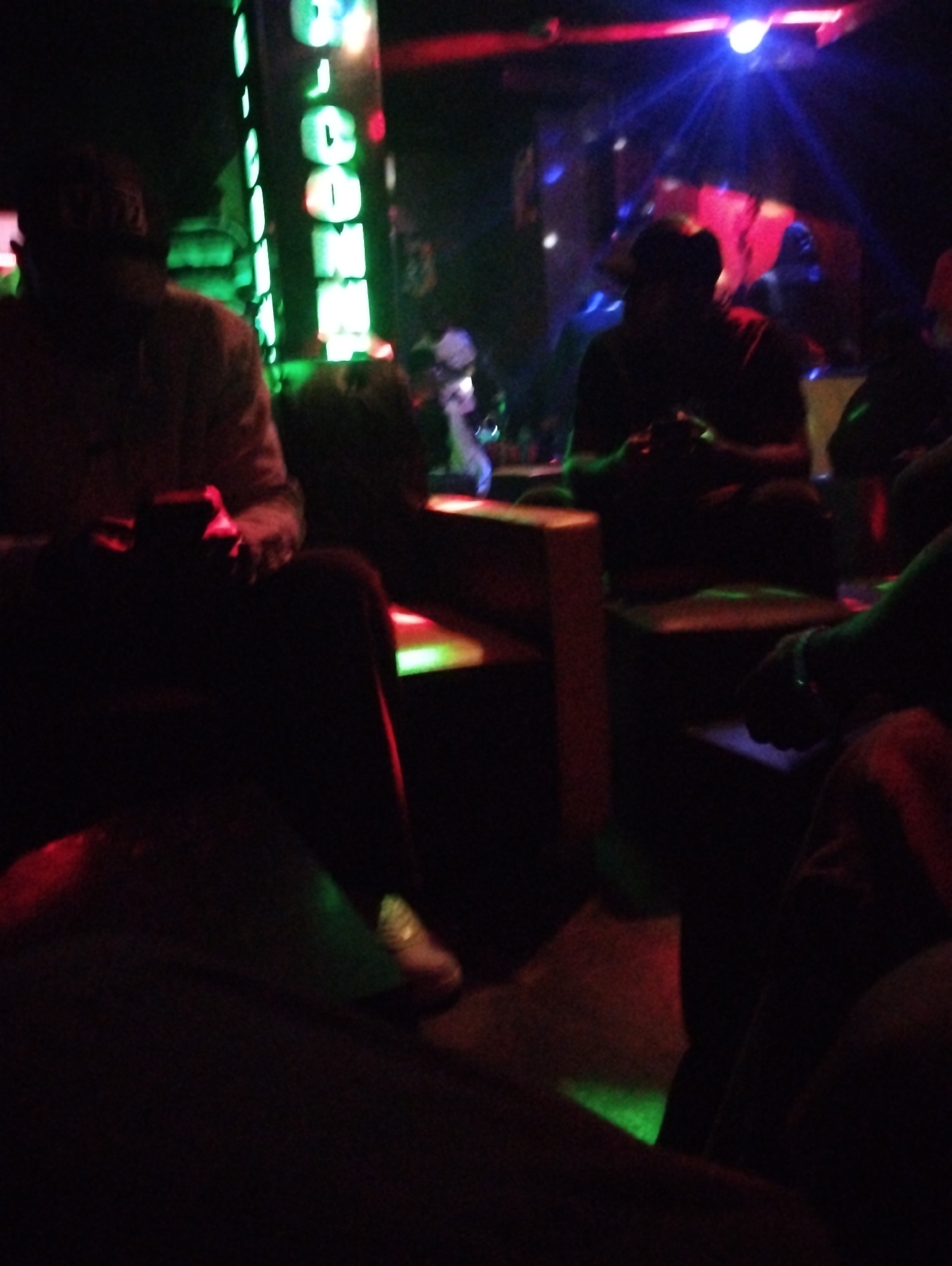 In a dimly lit indoor space in Gombe, Nigeria, several individuals are seated. The illumination comes from various sources, including green neon lights and a red light overhead. One person, wearing a hat and light-colored clothing, sits with their legs crossed, holding an object. Other individuals are also seated, some appearing to hold objects as well. The furniture appears to be wooden. The overall environment suggests a bar or nightclub setting with low lighting and possibly entertainment.