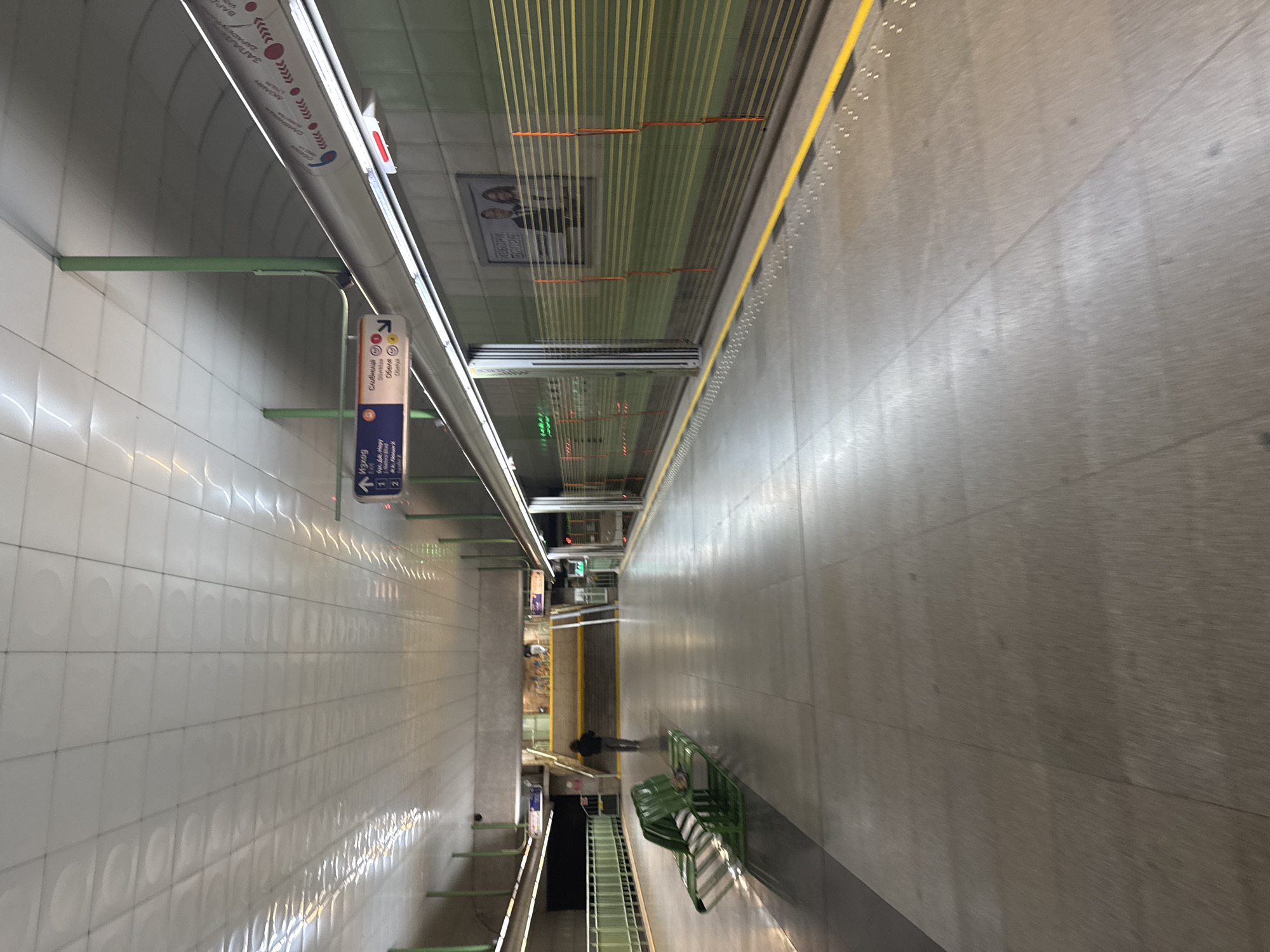The image depicts a subway station platform in Sofia, Bulgaria. The walls are tiled in white on one side and clad in gray on the other. Yellow tactile paving runs along the edge of the platform. Overhead, a network of yellow safety lines stretches above the platform. A sign indicates directions to Serdika metro station. A lone person is standing on the platform. Green benches are also visible.