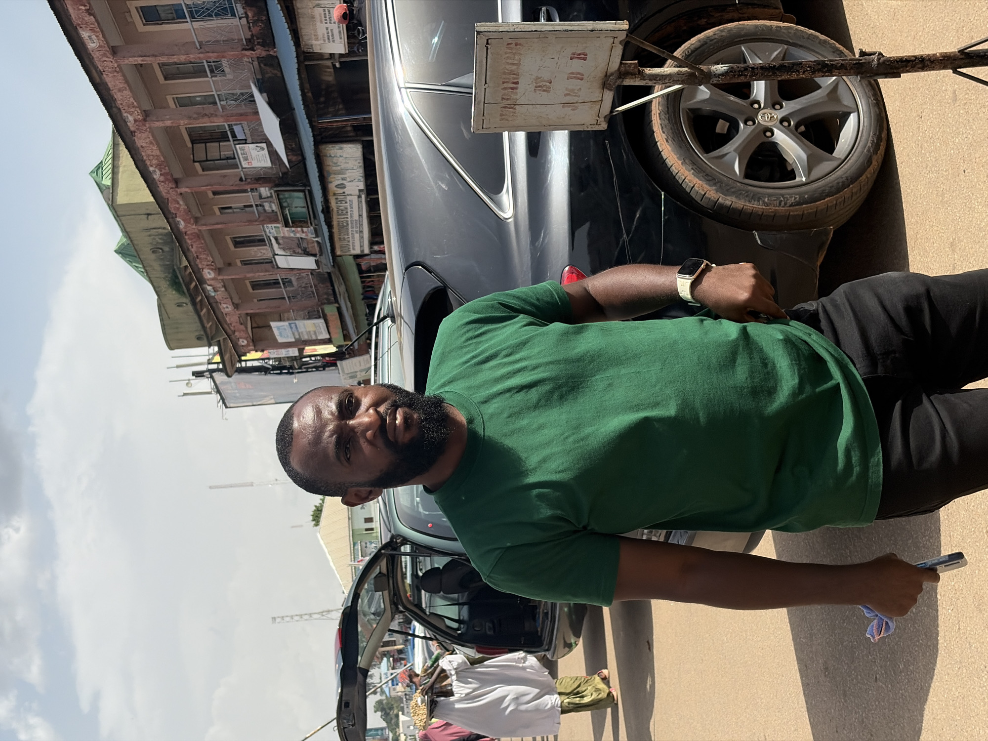 The image depicts a street scene in Jos, Nigeria. In the foreground, a Black man with a beard and a green t-shirt stands next to a dark-colored SUV. He is holding a light blue cloth and a smartphone. The SUV has its rear liftgate open, revealing a partially visible interior. In the background, a person wearing a white garment and a green headscarf, carrying a tray of items on their head, walks away from the camera. Further in the background, several buildings line the street, with some displaying signs and advertisements. A parking sign is visible in front of the SUV's wheel. The environment suggests an outdoor market or urban commercial area with a mix of vehicles and pedestrians. The sky is partly cloudy.