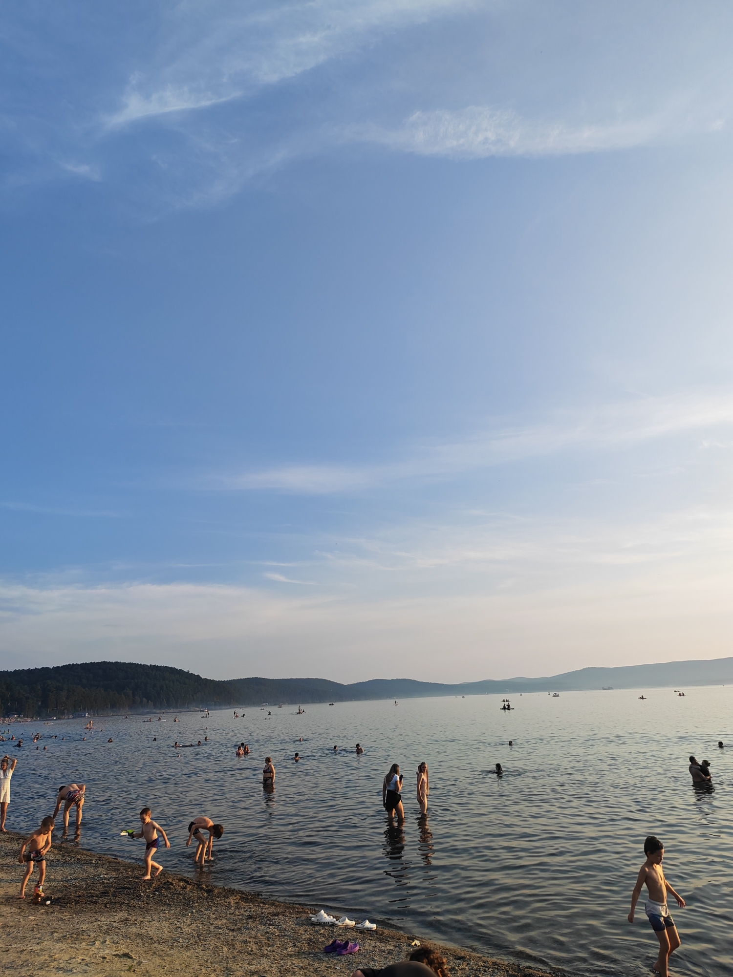 A wide shot captures a crowded beach and lake scene on a clear, sunny day. The sky is a bright blue with scattered wispy white clouds. In the foreground, people are wading in the shallow water near the shore, which is a mix of sand and pebbles. Several children are visible playing on the beach and in the water, some with toys. Further out in the lake, more people are swimming and boating. In the background, a tree-covered hill slopes down to the water's edge, and in the far distance, hazy mountains form the horizon. The overall atmosphere suggests a popular recreational spot, likely the Turgoyak beach in Turgoyak, Russia, given the characteristic landscape.