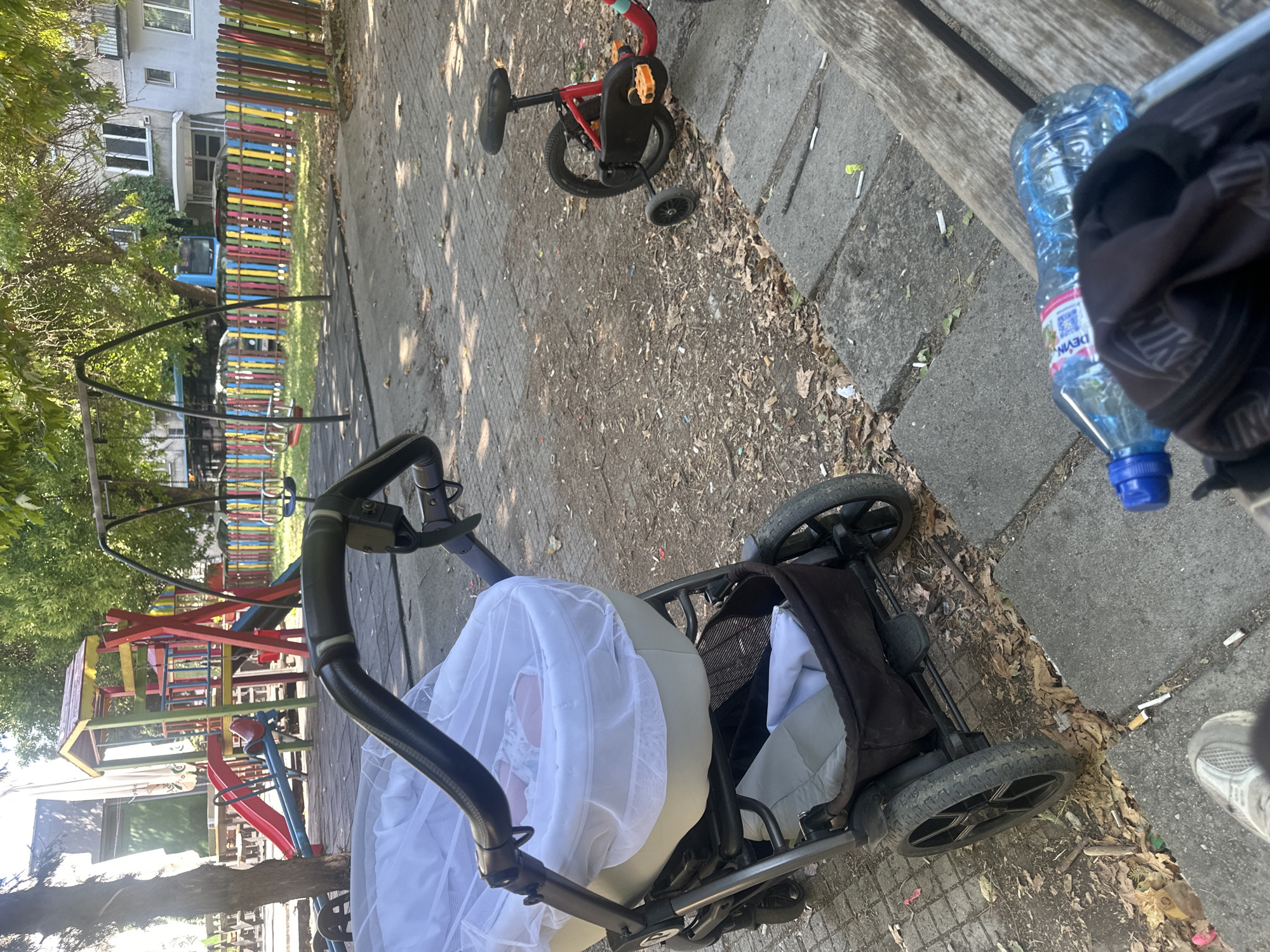 The image depicts a playground area in the city of Plovdiv, Bulgaria. In the foreground, a baby stroller is positioned, covered with a white mosquito net. A small red bicycle with training wheels is also visible. The background features a colorful playground structure with swings, slides, and a seesaw. A fence made of vertically arranged, multi-colored wooden planks separates the playground from buildings in the distance. The ground is paved with interlocking concrete blocks and scattered with fallen leaves and debris. A wooden bench is present on the right side of the image, with a plastic water bottle and a black backpack lying on it. The scene appears to be an outdoor recreational space.
