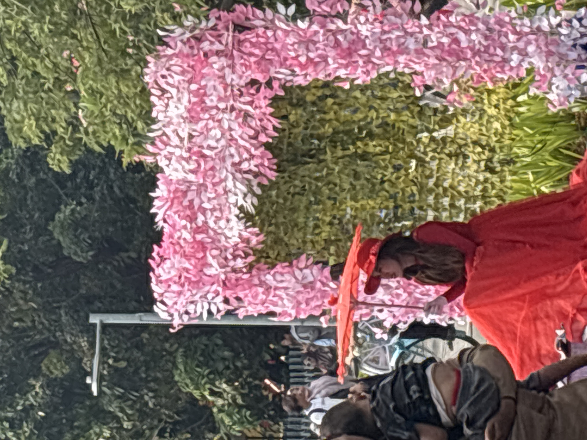 This is a close-up, eye-level, full-shot photograph of a woman in a red dress and hat, holding a red umbrella, in what appears to be an outdoor public space. She is seated on the right side of the frame, with her body angled slightly to the left and her gaze directed upwards and to the left. Behind her, to the left, is an archway adorned with pink and white artificial flowers, and behind that, a wall of green artificial foliage. In the background, slightly out of focus, are several people, a bicycle, and trees. The woman's dress is voluminous and red, and her hat is also red and adorned with a ribbon. The umbrella she holds is open and red. The environment suggests a decorative setup, possibly for an event or a photo opportunity. The presence of trees and a streetlamp in the background indicates an urban setting. The overall lighting suggests it is daytime. Based on visual cues and common event decorations in the area, this scene is likely from Taman Suropati in West Jakarta, Indonesia, a popular public park often used for events and gatherings.