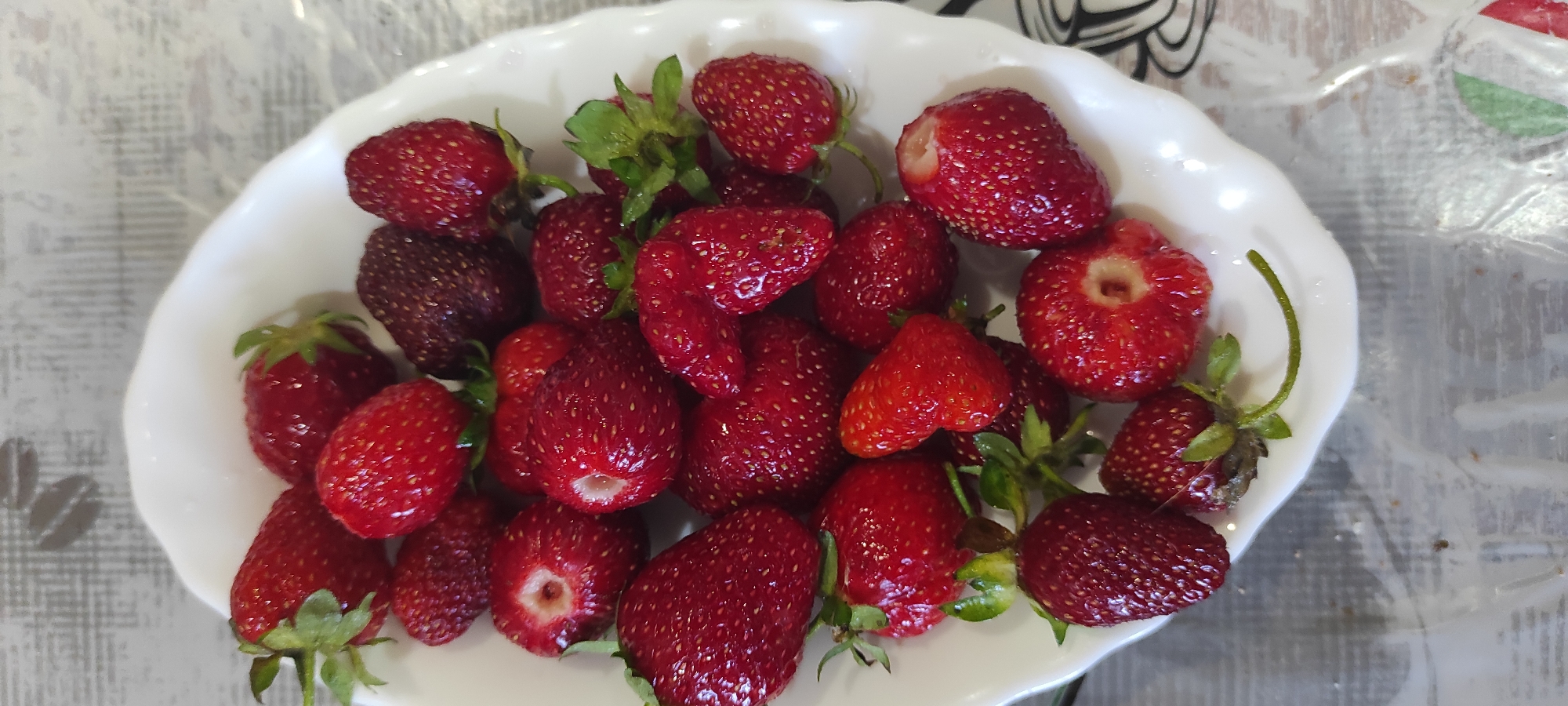 A white, fluted bowl overflowing with fresh strawberries sits on a table with a grey, patterned surface. The strawberries are a mix of vibrant red and a few darker, more mature ones. Some have their green stems and leaves attached, while others have been hulled. The lighting suggests an indoor setting, possibly a kitchen or dining area. The image was captured in Kobeliachok, Ukraine.