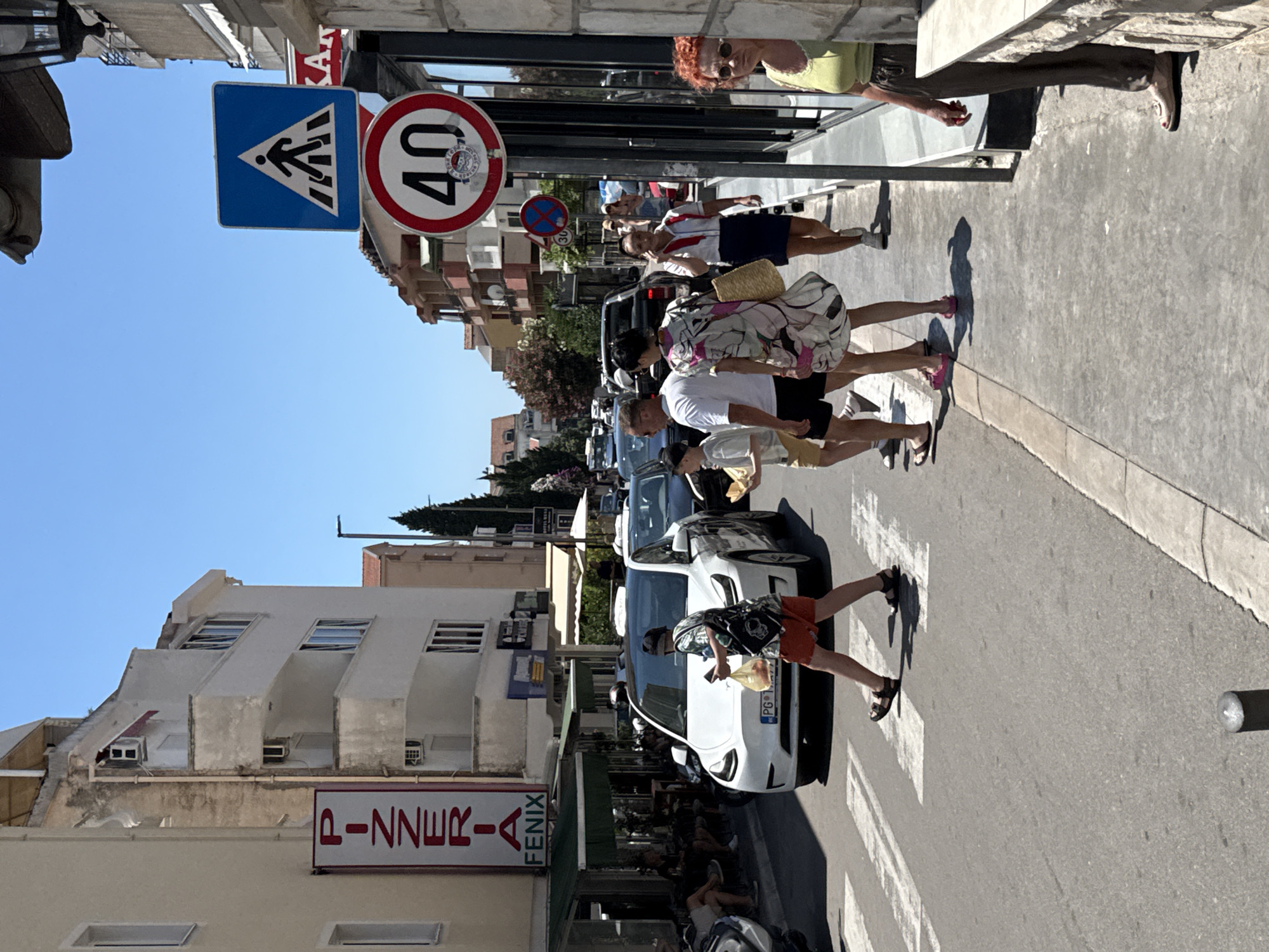 In Budva, Montenegro, on a sunny day, a white Tesla Model Y is stopped at a pedestrian crossing. A young person wearing orange shorts and a patterned t-shirt is crossing the street, holding a plastic bag. Behind the Tesla, a black SUV and other vehicles are visible on the street. On the sidewalk to the right, a woman with red hair is standing and looking towards the street. A tall signpost displays a blue pedestrian crossing sign and a speed limit sign with "40" inside a red circle. Further down the street, several people are walking on the sidewalk, and shops and buildings line both sides of the road. A vertical sign reading "PIZZERIA FENIX" is mounted on a building to the left.