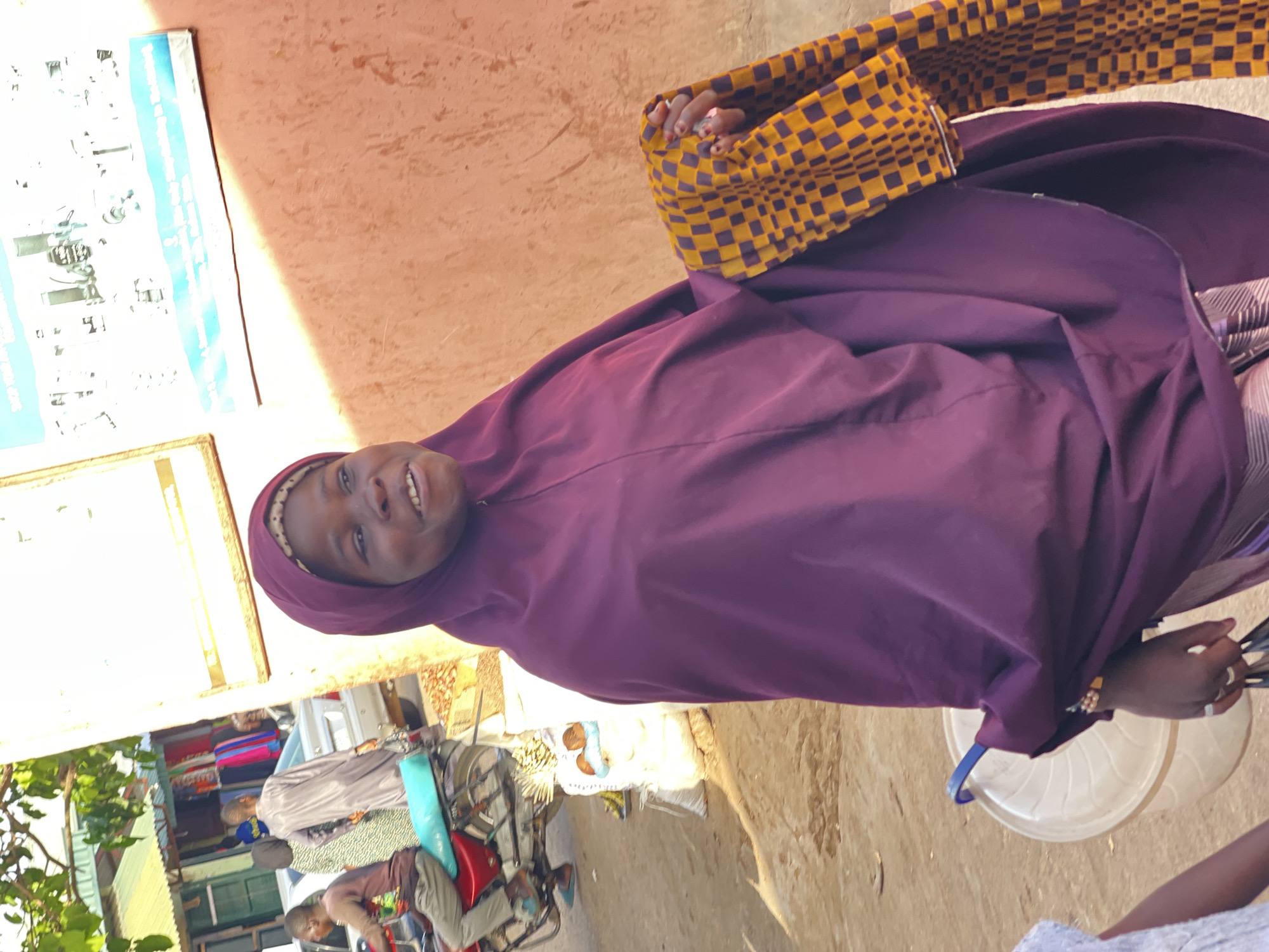 The image captures an outdoor scene in Samaru, Nigeria, illuminated by bright daylight, suggesting mid-day or afternoon. The main subject is a smiling woman in the foreground, wearing a modest purple hijab and a matching long dress. Her expression is friendly and open as she looks towards the camera.
Her hands are visible; one hand holds a distinctive yellow and dark purple checkered fabric, possibly a bag or head covering, while the other hand, adorned with red nail polish and a ring, rests near a light-colored plastic container on the ground.
In the background, a lively street or market environment unfolds. A person, likely a child or young man, is seated on a red motorcycle with blue accents in the lower left. Other blurred figures and buildings constructed with light brown or orange-toned walls are visible, depicting everyday activity.
On the wall behind the woman, two posters are displayed. The upper poster, with a blue border, has partially readable text, including phrases like "building a better future" and "Discover something new today at..." The lower poster clearly shows the text "SHOP NOW" and "SHOP ONLINE," indicating commercial advertisements. The overall setting appears to be a bustling local public space.