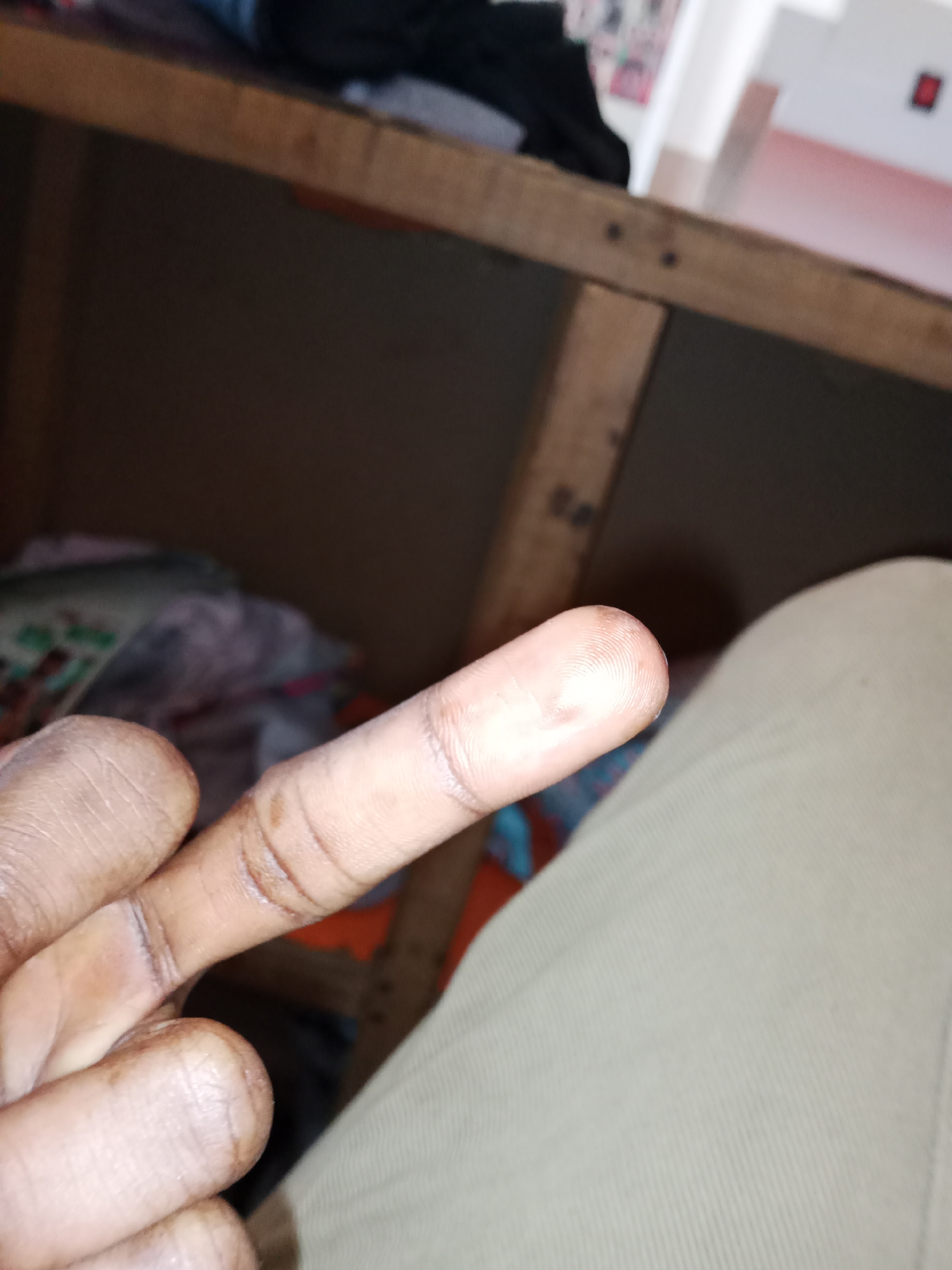 A close-up shot shows a person's hand and finger, with fingerprints visible on the fingertip. The hand is in focus and positioned in the foreground. In the background, a wooden shelf or structure is visible, with some blurred objects, possibly clothing or bags, placed on it. The setting appears to be indoors, possibly a room or storage area. The lighting suggests it could be daytime or well-lit indoors. There is no visible text in the image. The overall tone is neutral, with no specific emotions conveyed. The location context of Jalingo, Nigeria, provides a geographical reference.