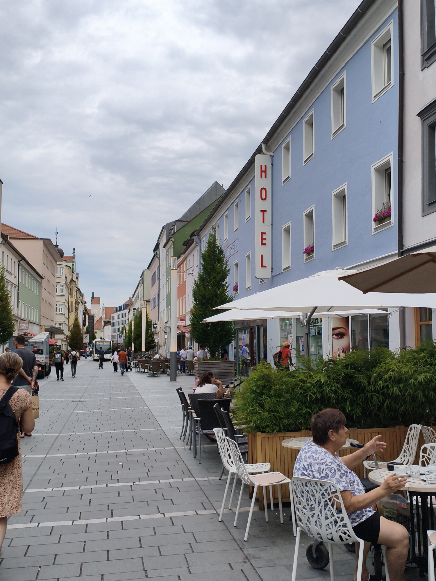 The image depicts a street scene in Regensburg, Germany, likely a pedestrianized zone. A blue building on the right side features a prominent red "HOTEL" sign. In front of the building, there is an outdoor seating area for a cafe or restaurant with white umbrellas providing shade. Several people are seated at tables, and a woman in a patterned shirt is gesturing with her hands. The street is paved with gray tiles and lined with colorful buildings on both sides, typical of European city architecture. A bus is visible in the distance, and various people are walking along the street. Lush green bushes border the outdoor seating area. The sky is overcast.