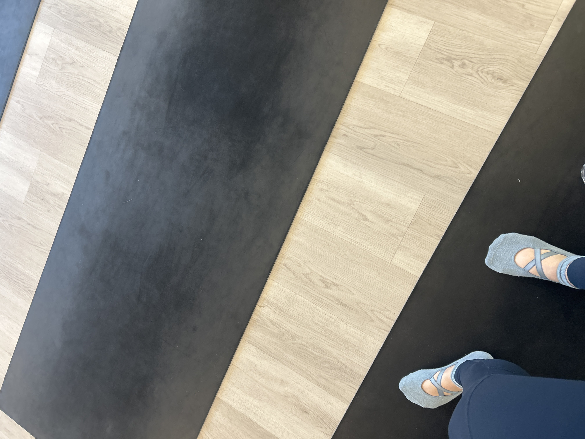 A top-down shot features a person's feet in light blue ballet-style socks on a black yoga mat, surrounded by a light wood-tone floor. The image shows parts of three yoga mats arranged parallel to each other, alternating with strips of the light wood floor. The feet are positioned on the mat to the right. The image suggests an indoor setting likely in a studio designed for yoga or fitness activities. No city is visible in the image.