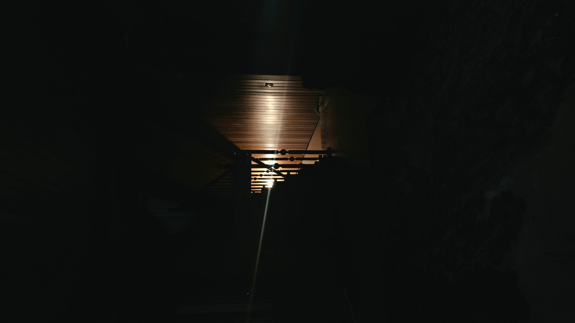 The image depicts a dimly lit interior scene, likely a residential hallway or staircase. A beam of light, possibly from a flashlight or headlights, cuts across the frame from the left, illuminating a wooden-paneled wall and the railings of a staircase. The staircase itself has turned wooden spindles and is partially obscured by darkness. The overall atmosphere is one of shadow and selective illumination, with the light source casting long shadows and highlighting the textures of the wood. There are no individuals visible in the frame. The environment appears to be indoors, with a slatted ceiling visible in the upper portion of the image. There is no indication of the city of Il'inskiy, Russia within the image itself.