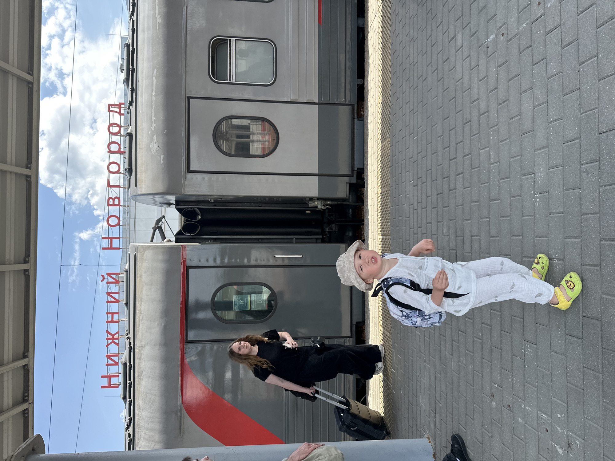 A young child and woman stand on a train platform in Nizhny Novgorod, Russia, next to a passenger train. The train has a red stripe and a large red sign that reads "Нижний Новгород" (Nizhny Novgorod). The child wears a light-colored outfit, a hat, a backpack, and yellow sandals, and appears to be looking at the camera. The woman, standing behind the child, wears dark clothing and pulls a black suitcase with wheels. The platform is paved with gray rectangular bricks. Overhead, a partial view of a canopy and some sky with clouds can be seen. Other people's legs and shoes are visible in the bottom part of the frame.