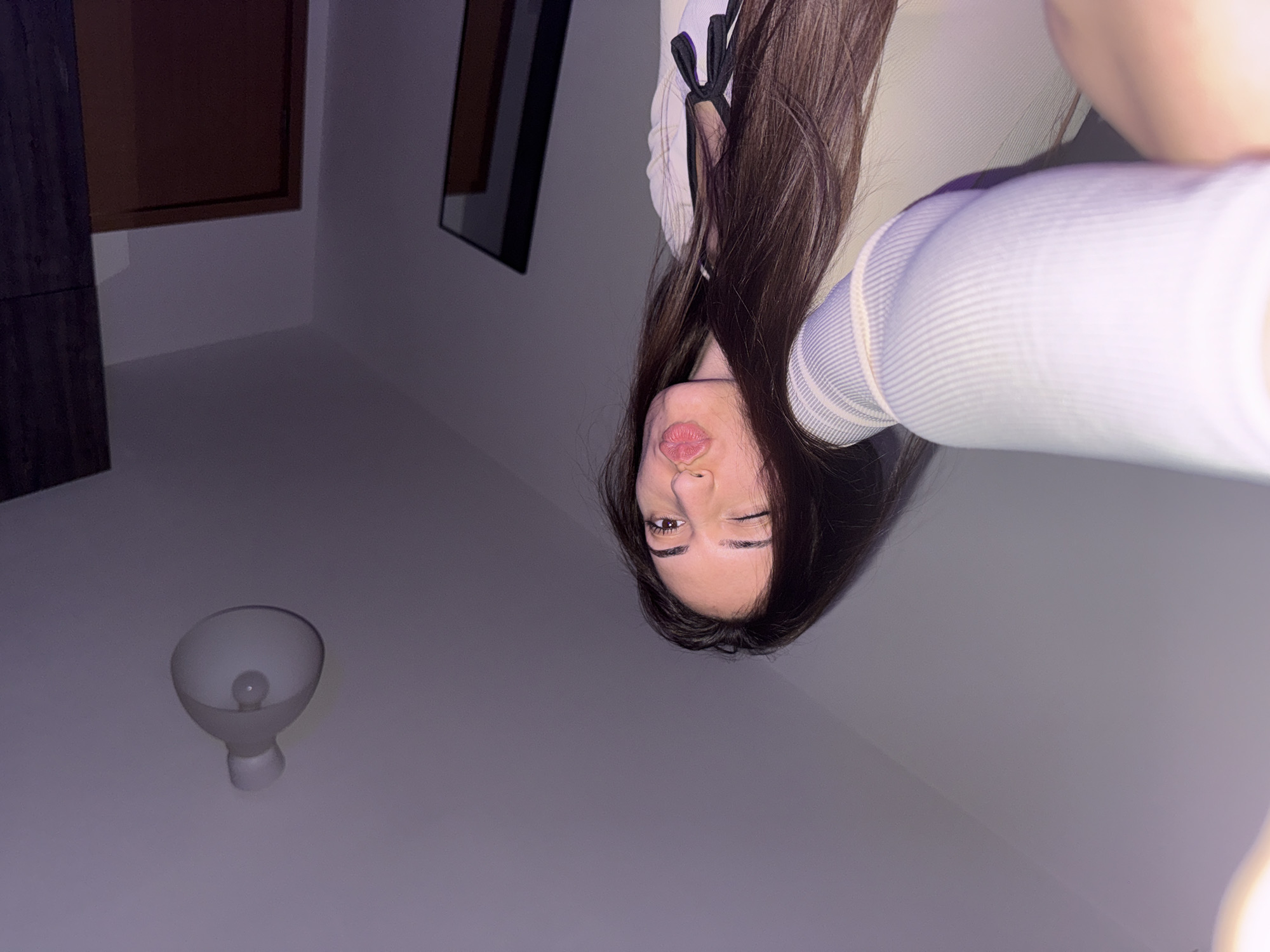 The image depicts a female individual taking a selfie indoors in Plovdiv, Bulgaria. The individual, positioned centrally, has long, dark hair and is winking her left eye while pursing her lips. Her right arm is extended forward, holding the camera, with a light-colored, ribbed long-sleeved garment visible. She wears this over a darker top featuring a small bow detail at the neckline. The background features light-colored walls and ceiling. An illuminated, white, bell-shaped ceiling light fixture is mounted on the ceiling in the upper right quadrant. To the right, a dark wooden wardrobe or cabinet is partially visible, alongside a dark-framed mirror or reflective object and a brown door within a light-colored frame. The scene is illuminated by artificial light.