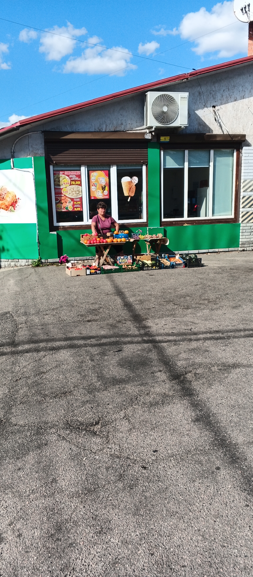 In Smila, Ukraine, a woman stands behind a table displaying various fruits and vegetables. The table is set up in front of a green-colored storefront with large windows. The woman wears a purple shirt. Piles of produce, including tomatoes, oranges, and grapes, are arranged on the table and in crates on the ground. In the background, the storefront windows feature advertisements for discounts and images of ice cream. An air conditioning unit is mounted on the exterior wall above the storefront. The scene takes place outdoors on an asphalt surface.