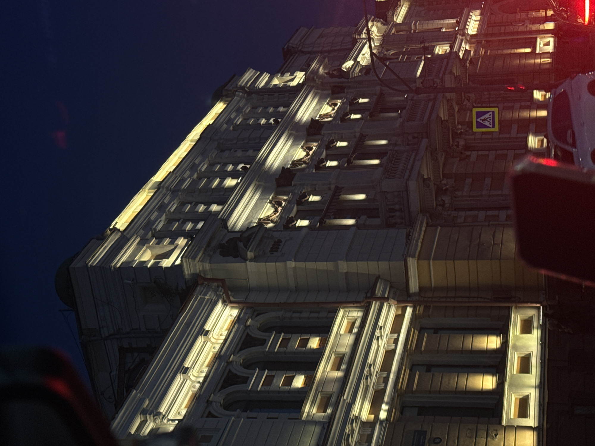 A nighttime, low-angle urban street scene captures a multi-story, ornate building facade illuminated by warm white upward-facing architectural lights. The building, constructed of a light-colored material, features a prominent cornice line along the roof and various decorative elements including pilasters, arched windows, recessed rectangular windows, and several balconies with elaborate railings. An inscription "1896" is visible on the upper-middle section of the building facade. A reddish-brown drainpipe runs vertically down the left side of the building. The upper-left corner of the image is partially obscured by dark, blurred material, possibly from within a vehicle, while a faint dark green dome is visible on the building's roof against the dark blue sky.
In the mid-ground, a dark street light pole with a curved arm stands in front of the building. A yellow and blue rectangular pedestrian crossing sign is attached to a pole, clearly visible against the building's illuminated facade.
The foreground is dominated by the interior of a vehicle, with blurred dark and red shapes partially obscuring the bottom of the scene. Visible on the street are parts of two vehicles: a white car (possibly an SUV) is seen from the front-left, facing right, with its headlights off, and the red taillight of another vehicle is visible on the far right. Shadows and indistinct forms that may be pedestrians are vaguely visible at street level near the white car and the pedestrian crossing. The sky overhead is a dark navy blue, with a subtle reddish-purple glow along the horizon on the far right.